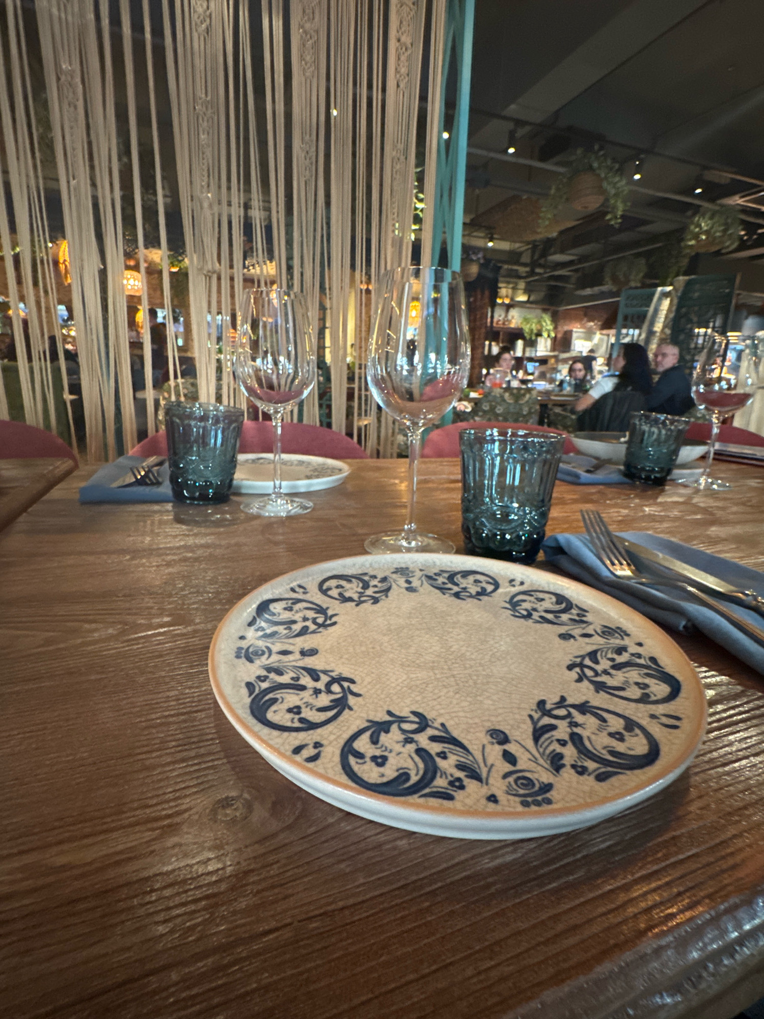 A close-up, slightly angled, eye-level shot shows a table setting at a restaurant in Ufa, Russia. The table is made of dark wood with a prominent grain. In the foreground, a decorative ceramic plate with a blue floral pattern is positioned slightly off-center to the right. To its left, a folded blue napkin with a fork and knife placed on top is visible. A dark blue glass tumbler stands next to the napkin. In the background, behind the table, are two wine glasses, one on either side of a similar blue tumbler. Beyond the table setting, a dimly lit restaurant interior is visible. White macrame strings hang vertically, obscuring parts of the background. Several patrons are seated at tables in the distance, and plants are suspended from the ceiling, indicating a possibly botanical-themed ambiance. The overall lighting is subdued, creating a warm and intimate atmosphere.