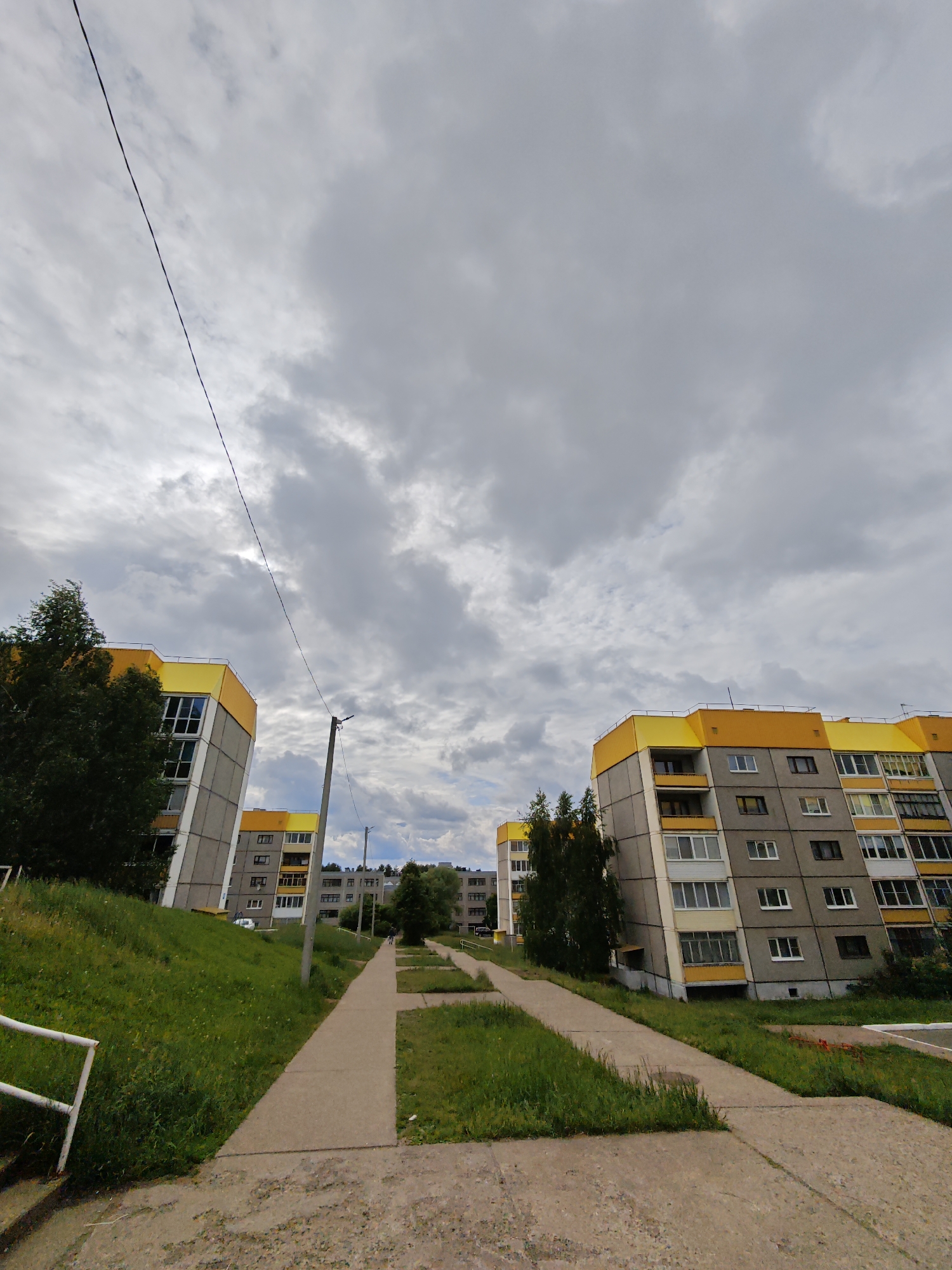 The image captures a residential landscape in Markovskiy, Russia, under an overcast daytime sky. Dominating the scene are several multi-story apartment buildings, characterized by their grey facades and distinctive bright yellow top floors and roofs. These buildings feature numerous windows and some balconies. Green grassy areas, some on slopes, surround the buildings and flank a winding concrete pedestrian path that extends into the distance. Scattered trees, both large and small, are interspersed throughout the green spaces. In the foreground, a dark utility pole stands beside the path, with a thin power line stretching diagonally across the cloudy sky. A white metal railing is partially visible in the lower left. The heavy, grey clouds suggest gloomy weather or an impending storm. The overall impression is a functional, albeit somewhat stark, urban environment with established green spaces. No people or visible text are present.