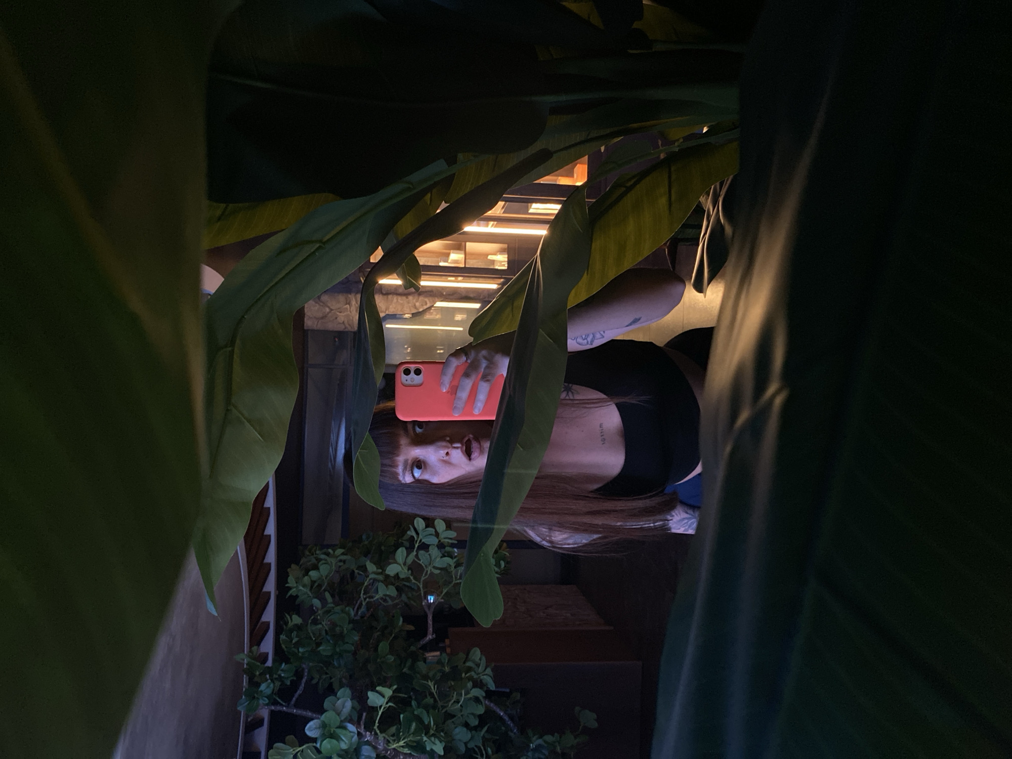 A female is taking a selfie with a smartphone, framed by large green leaves of a plant. She is wearing a black top and has brown hair with bangs. There is a small potted plant to the left. A building facade with lights is visible in the background. The location is Votkinsk, Russia.