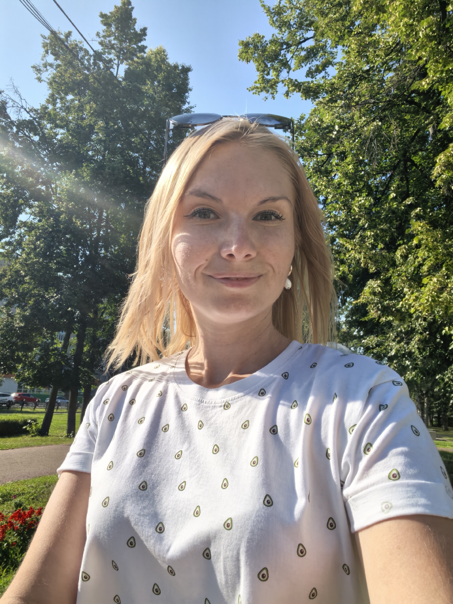 This is a selfie of a blonde woman taken outdoors in Sterlitamak, Russia, on a sunny day. She is wearing a white t-shirt with a repeating avocado pattern and large white earrings. The background consists of lush green trees and a clear blue sky, with power lines visible above. Sunlight filters through the leaves, creating a dappled effect on the woman and her surroundings. In the distance, a road with parked cars and a sidewalk can be seen, suggesting a park or residential area within the city. The woman is looking directly at the camera with a slight smile.