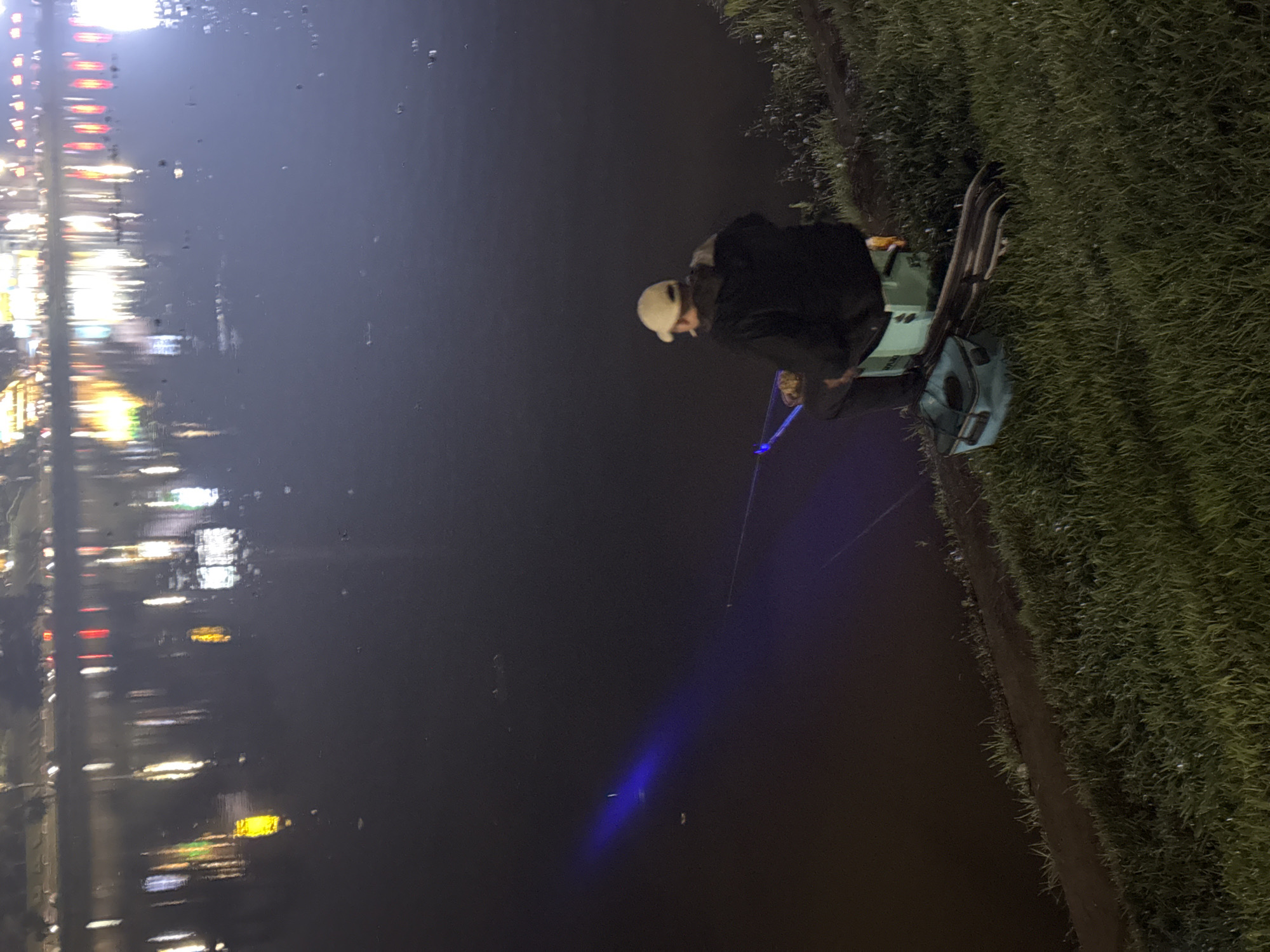 The image depicts a person fishing at night in Da Lat, Vietnam. The individual is wearing a light-colored cap and a dark jacket, seated on a chair next to fishing gear. A fishing rod is visible, extending towards the water, with a faint blue glow emanating from near the water surface, presumably from the line. The water reflects the blurred lights of the city, indicating an urban setting. The adjacent shoreline is covered in grass.