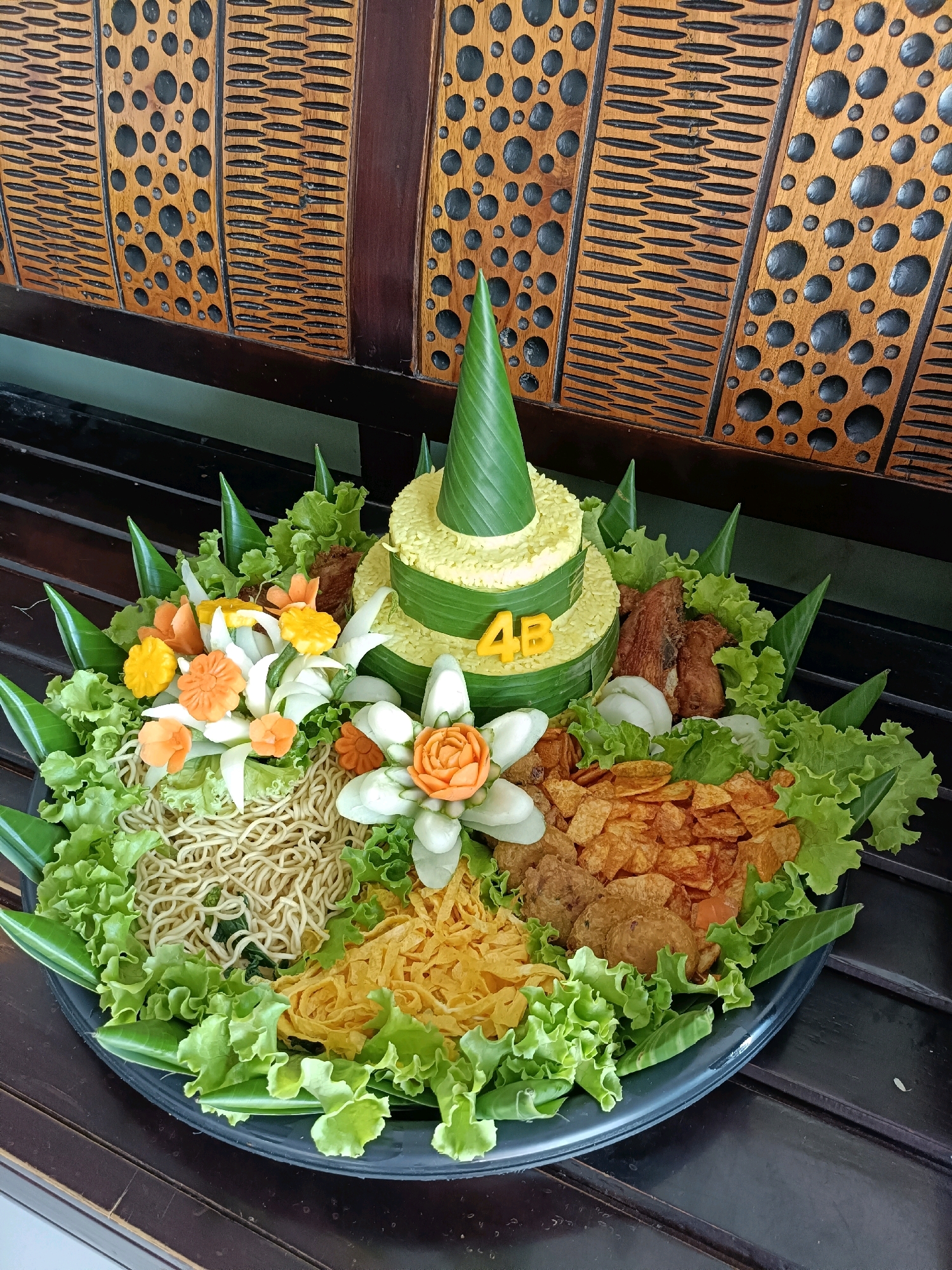 A conical-shaped yellow rice dish, known as a tumpeng, is presented on a dark circular platter, adorned with various food items and decorative elements. The tumpeng, layered with banana leaves, is topped with a green cone made from a banana leaf and features yellow letter cutouts spelling "4B" on its side. Surrounding the tumpeng are an assortment of ingredients, including shredded noodles, scrambled egg strips, fried crispy snacks, fried chicken pieces, and decorative carved vegetables resembling flowers in orange and white. The platter is further embellished with fresh lettuce leaves and more banana leaf cones. The tumpeng and platter are placed on a dark wooden surface, which appears to be a table or bench. The background consists of a wooden wall with an intricate carved pattern of circular and oval shapes, set against a dimly lit interior. The lighting is warm and appears to be natural, casting soft shadows. This scene is located in Tamanan, Indonesia.