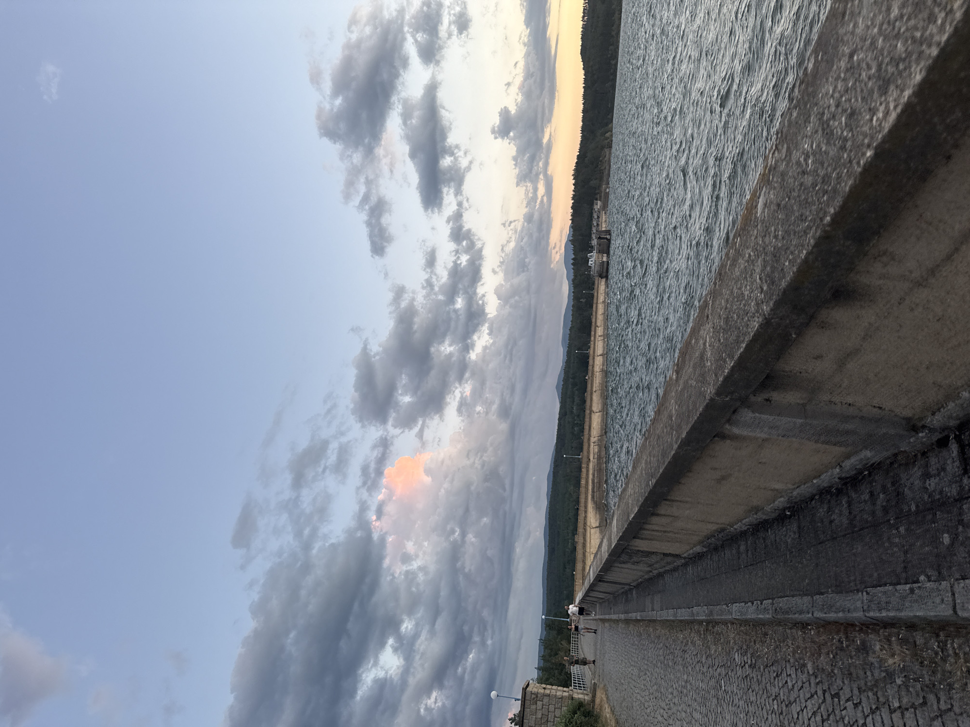 The image shows a dam at Koprinka, Bulgaria, on a cloudy day. A paved walkway runs along the top of the dam, with a low concrete wall separating it from the water. A few people are walking on the walkway. The water is choppy, with small waves. The sky is a mix of blue and gray clouds, with a patch of pink light from the setting sun visible in the distance. A line of trees borders the far side of the reservoir.
