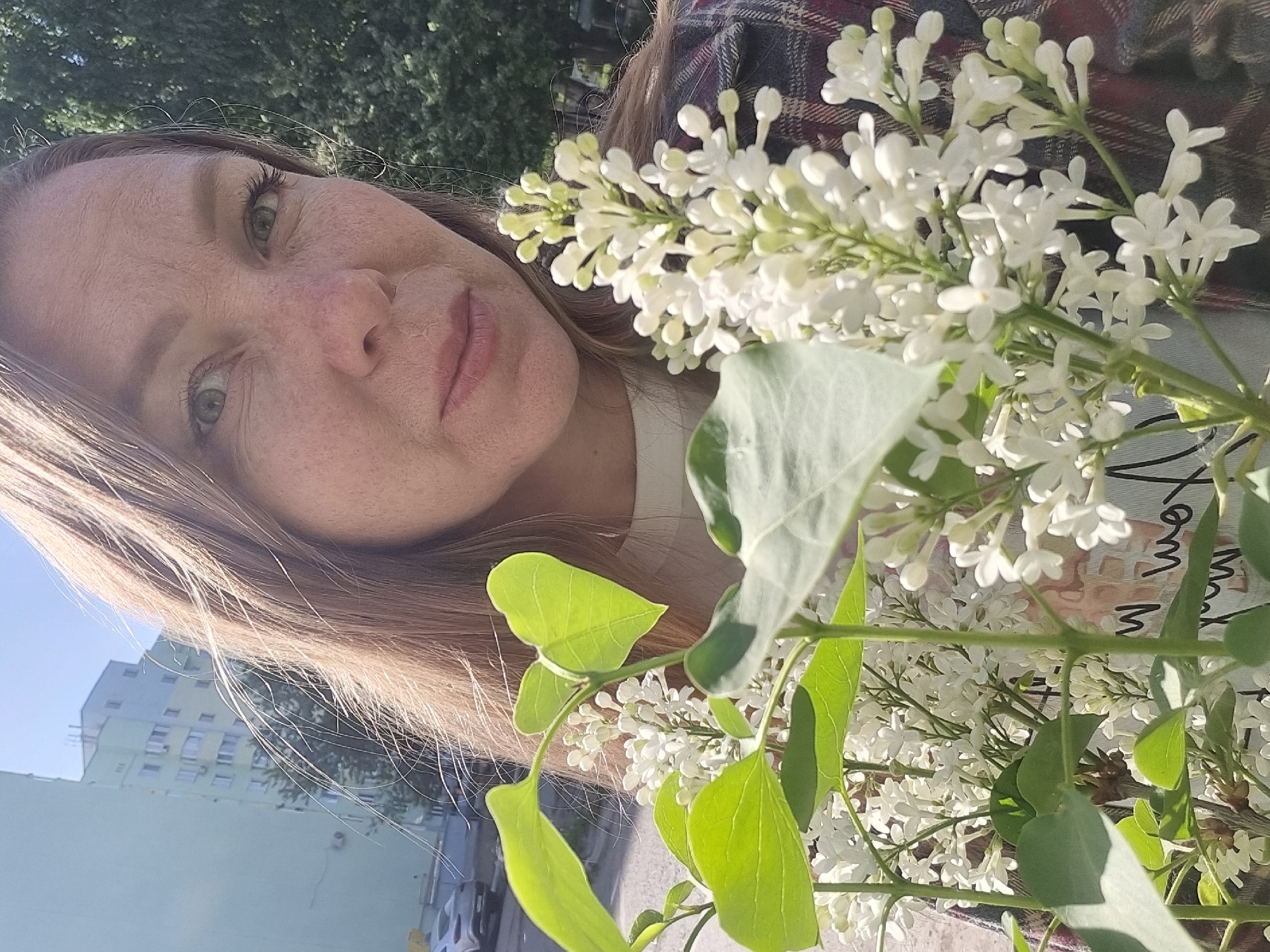 A fair-skinned woman with freckles and light brown hair is the central figure, holding a bouquet of white lilacs. She is looking at the camera with a neutral expression. Her clothing is partially visible, including what appears to be a plaid shirt. The lilacs have bright green leaves and clusters of white flowers. In the background, there is a light green apartment building, a common architectural style in Chaikovsky, Russia. The overall composition suggests the photo was taken outdoors on a sunny day.