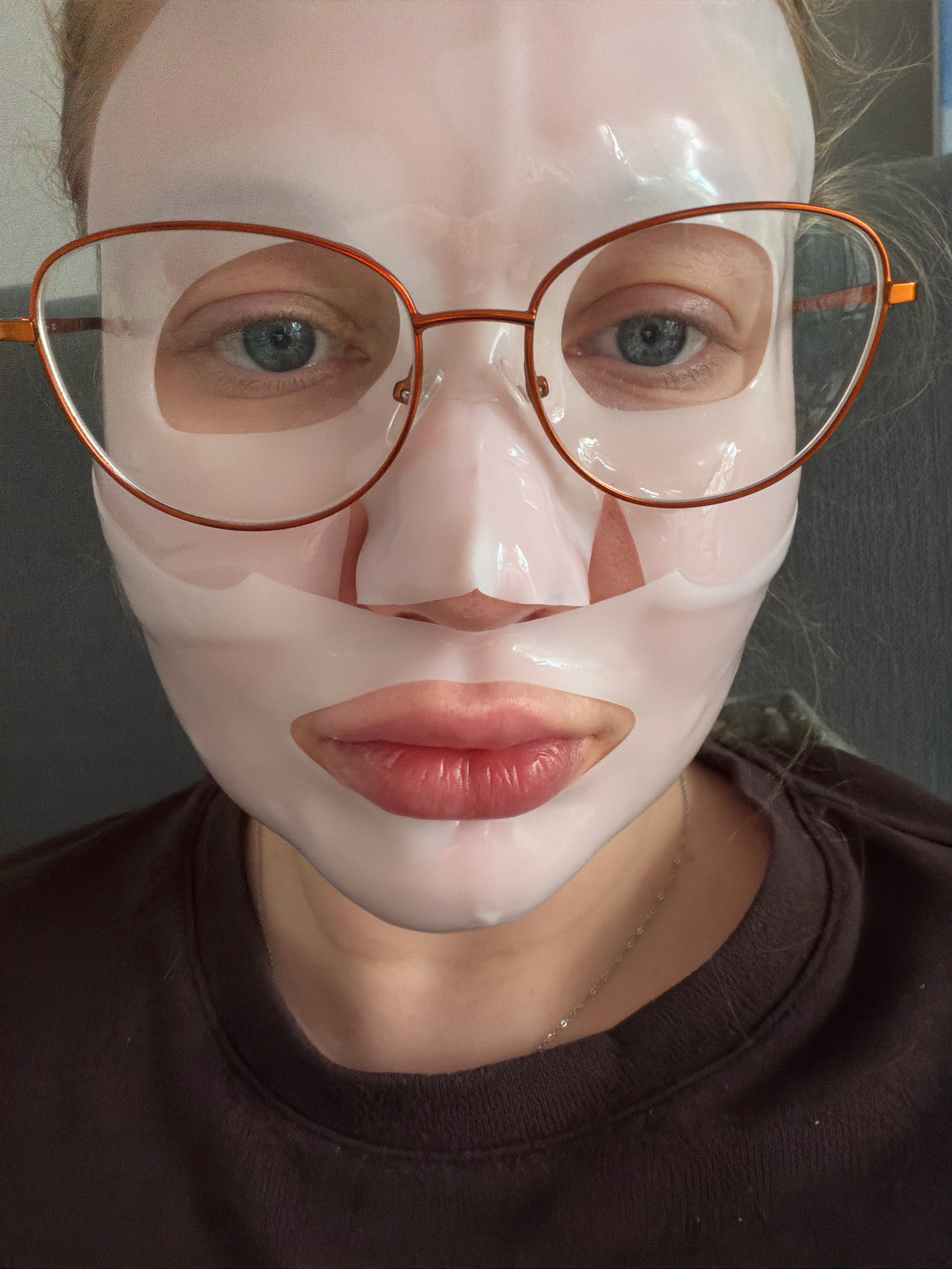 A close-up, front-facing, head-and-shoulders shot of a person wearing a white sheet mask and orange-rimmed glasses. The mask covers the entire face, with cutouts for the eyes and mouth. The person's eyes are visible and are blue. The lips are also visible through the mask and appear to have a pinkish-red tint. The person is wearing a dark brown crew-neck sweatshirt. The background is blurred and appears to be a neutral, indoor setting. There is no information in the image to determine the location in the city of Koszalin, Poland.