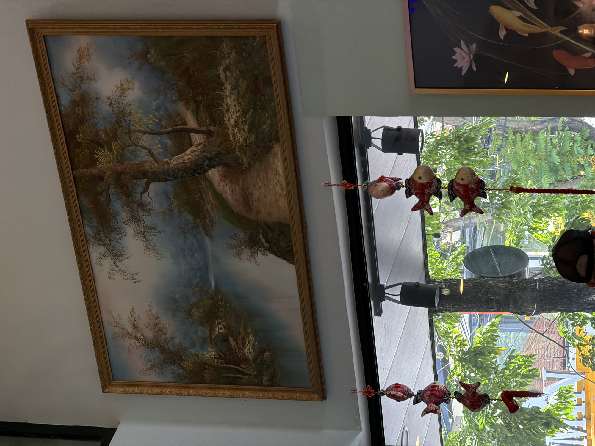 The image shows a room interior with two framed pictures hanging on the wall. One is a landscape painting with a river and trees, and the other depicts koi fish. A string of decorative fish is hung in front of a window. The window provides a view of the outside, displaying trees and buildings. The location is Đà Nẵng, Vietnam.