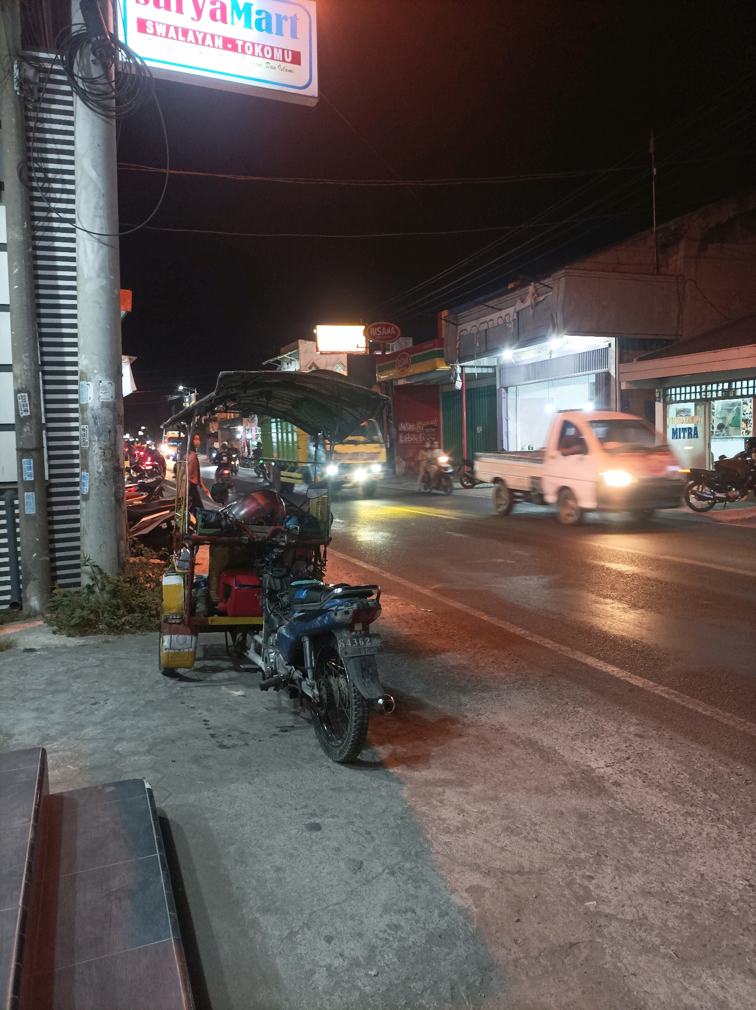 The image depicts a street scene at night in the city of Blimbing, Indonesia. On the left, a pole with electrical wires is prominent, beside it a sidewalk with a three-wheeled vehicle, likely a modified motorcycle or auto rickshaw, parked. This vehicle has a covered seating area and a blue motorcycle attached to its rear. Further down the sidewalk, there are some bushes. The street itself is wet, reflecting the lights from street lamps and passing vehicles, suggesting recent rain or the presence of puddles. Several motorcycles and a white pickup truck are visible on the road. The truck's headlights are illuminated and it appears to be in motion. Buildings line the right side of the street, with illuminated signs visible, including "SURYA Mart" and "MITRA". The overall atmosphere is that of a typical Indonesian street at night, with moderate traffic and commercial activity.