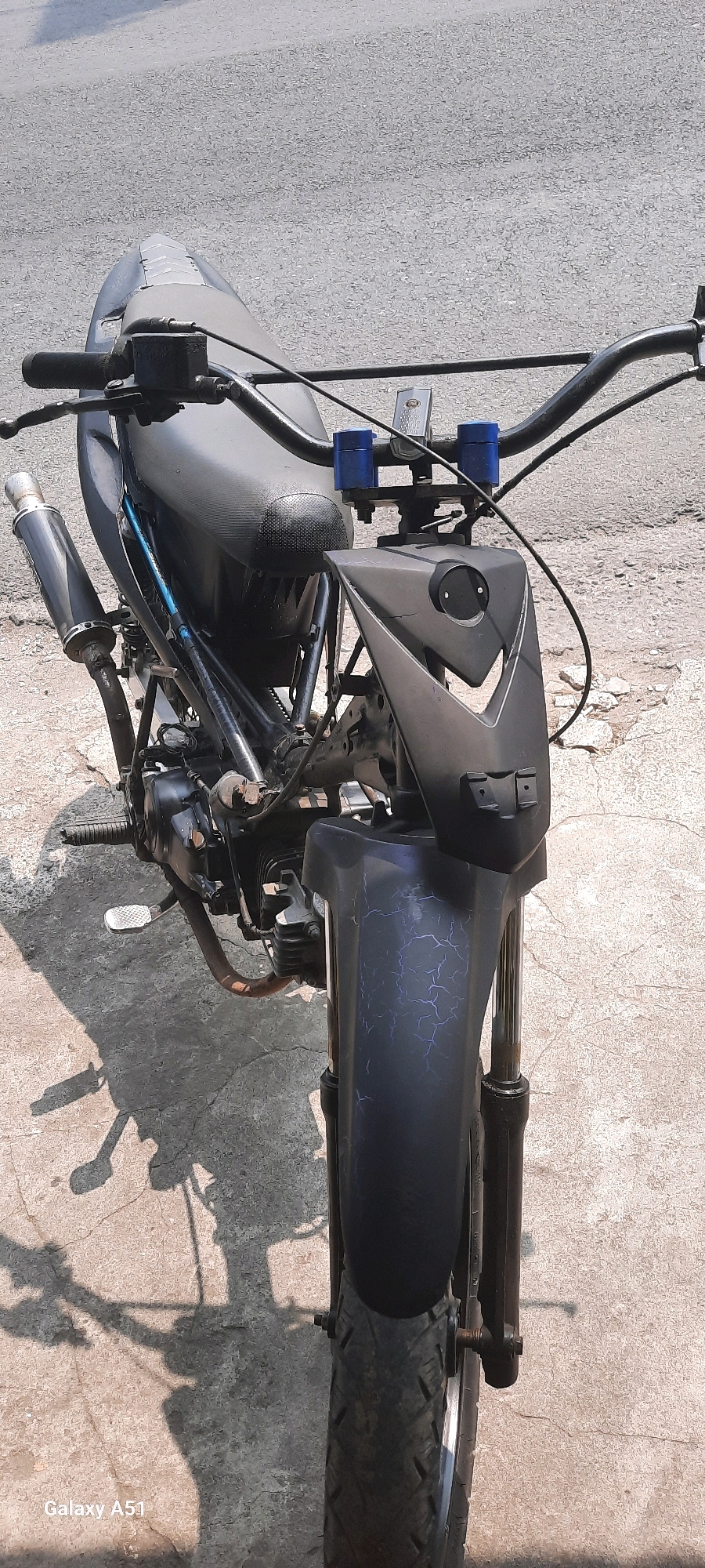 This is a medium shot, eye-level, outdoor, natural light, close-up photograph of a modified motorcycle. The motorcycle is dark gray with black accents and has a custom-designed front fairing that resembles a face with sharp angles and circular "eyes." The handlebars are raised and appear to have blue accents on the risers. The seat is black and textured. A black exhaust pipe is visible on the left side. The front fender has a crackled, dark blue paint design. The motorcycle is parked on a paved surface, likely a street or sidewalk, with some visible cracks and texture. The background consists of a gray, asphalt-like surface. The shadow of the motorcycle is cast on the ground to the left. The photograph is watermarked with "Galaxy A51" in the lower-left corner. The scene is likely located in the city of Tangkil, Indonesia, as indicated in the prompt.
