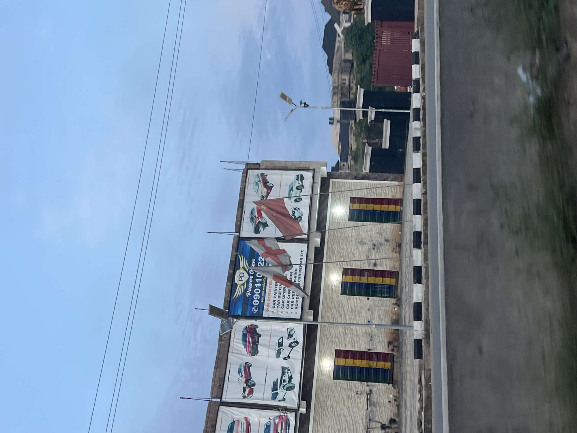 In Orifite, Nigeria, a medium-shot captures a building exterior under a twilight sky with scattered clouds and power lines. The building features a brick facade with prominent, brightly colored doors: red, yellow, and green. Above the doors, a large billboard displays various car models and advertisements for "Power Oven," listing services such as car painting, buffing, conditioning, and repair. Two flags are attached to poles in front of the billboard, one white with a red cross and another red flag. A modern street light stands near the building. To the right of the building, a dark-colored gate with pillars and a red metal fence is visible, with a lion statue positioned on the right side of the frame, partially obscured. A paved road with black and white markings runs in the foreground.