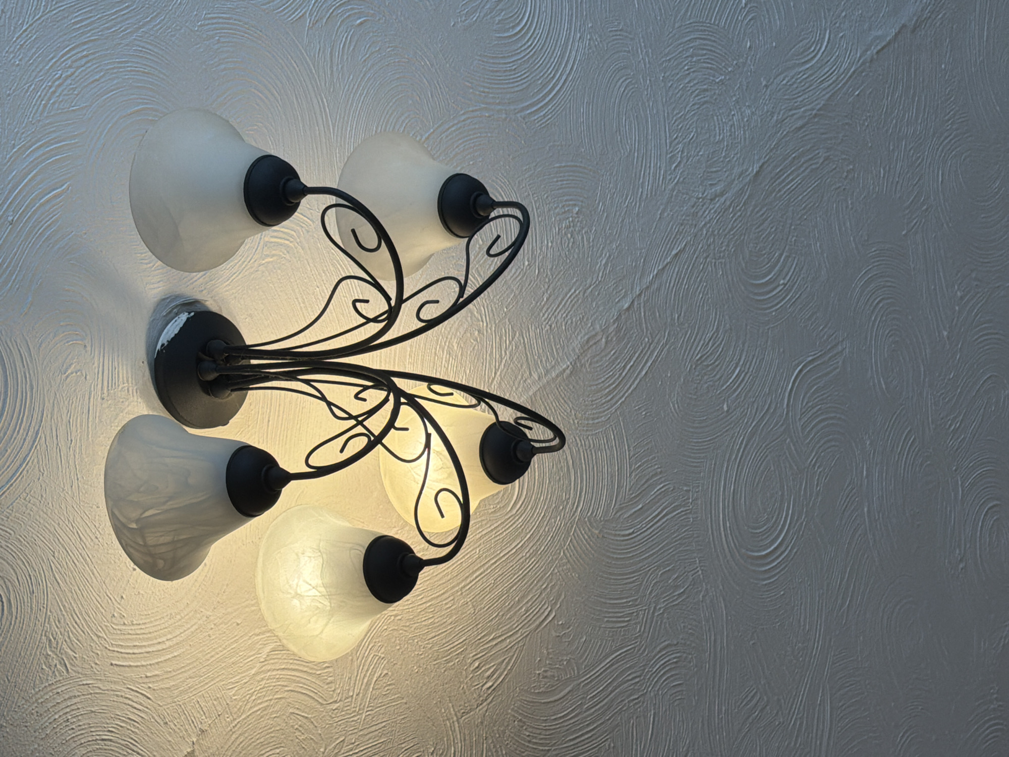 A five-bulb ceiling light fixture dominates the left side of the frame, casting light onto a textured, swirling-patterned wall. The fixture is black metal with white, tulip-shaped glass shades, and the bulbs are illuminated. The wall is a light grey color. The image lacks contextual clues regarding a specific location within Bearpark, United Kingdom. There are no people or other discernible subjects or actions in the frame.