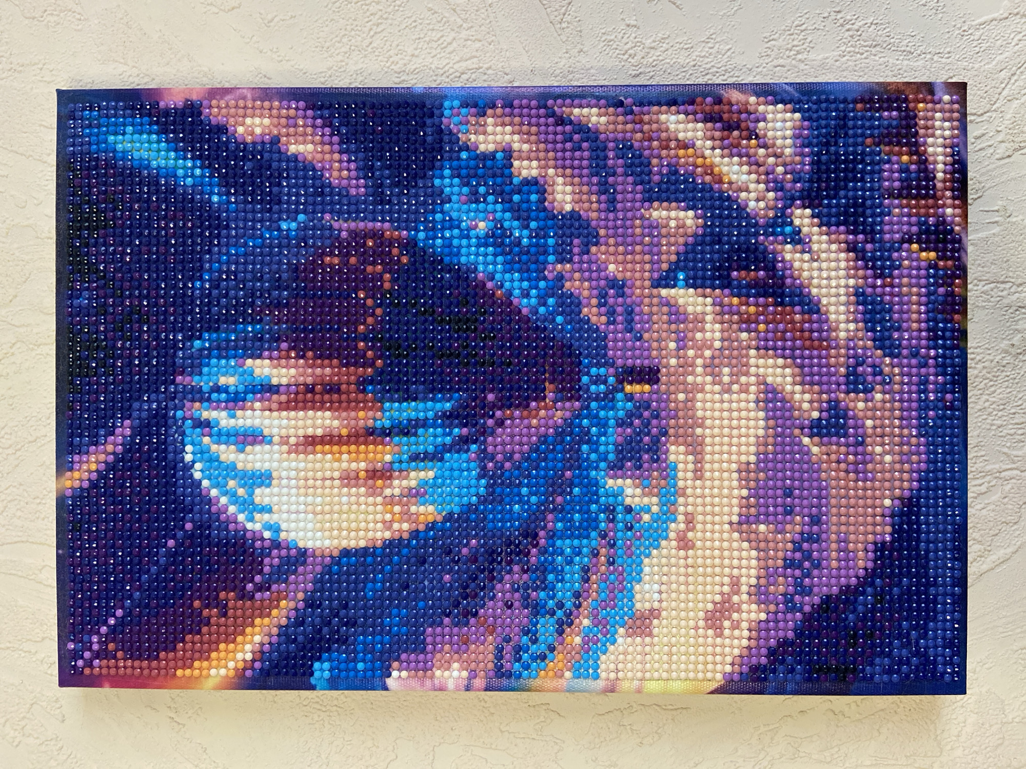 A rectangular diamond painting is displayed against a textured off-white wall. The painting depicts an abstract image dominated by shades of blue, purple, orange, and light beige. The painting is densely covered with small, round, and multi-faceted diamond-like pieces. The painting appears to be mounted on a wooden frame. There is no information present in this image to suggest this image was taken in Chaikovsky, Russia.
