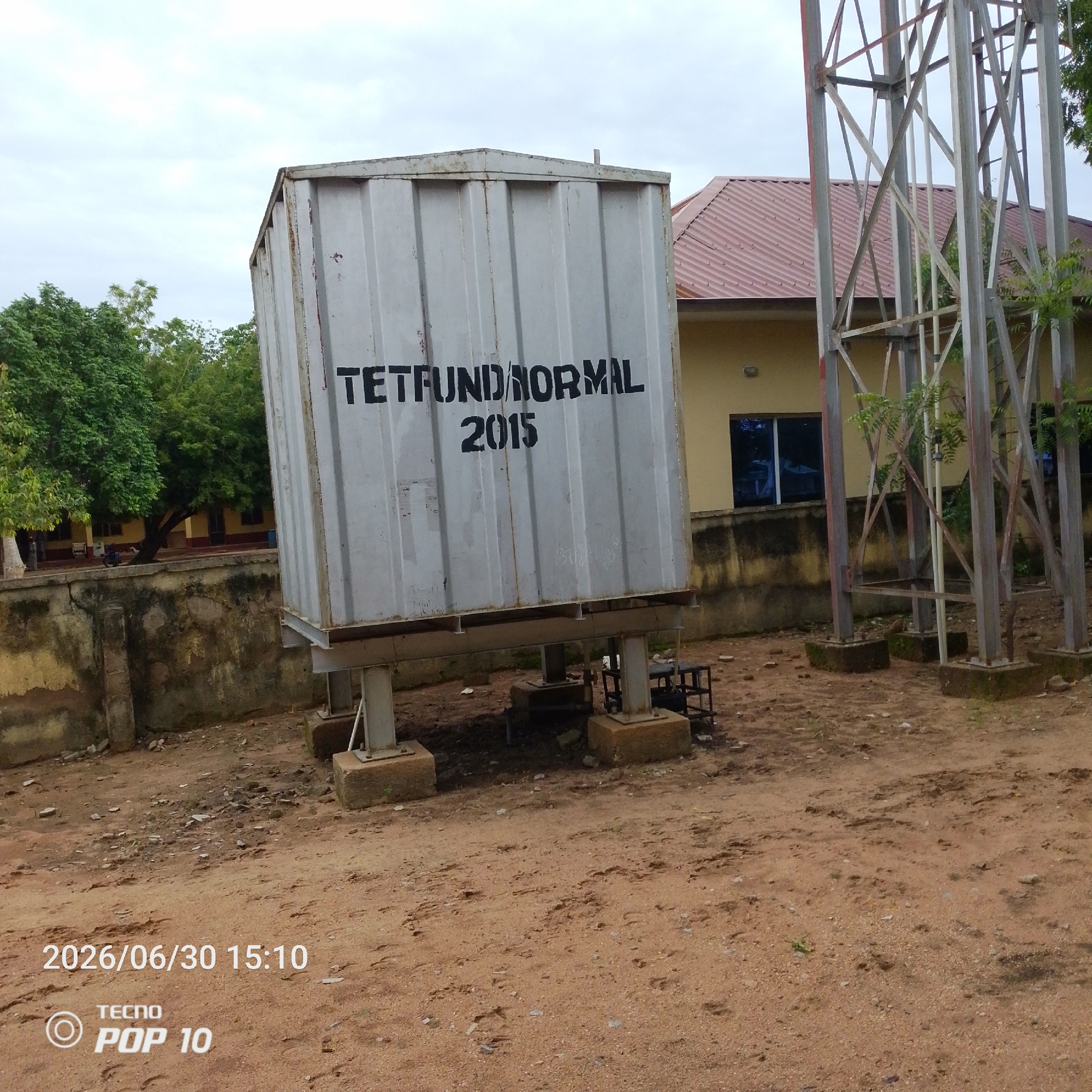 The image depicts an outdoor scene with a large, corrugated metal water tank prominently displayed. The tank is silver and has black lettering that reads "TETFUND NORMAL 2015." It is elevated on four metal legs, which are anchored to concrete bases. To the right of the water tank is a tall, lattice-like metal tower, likely a communication or utility tower, with metal beams and supports.
In the background, a light yellow building with a reddish-brown, ridged roof is visible. Several windows with dark panes are apparent on the building's facade. Green trees with dense foliage surround the area, suggesting a natural or park-like setting, though the ground is covered in dirt and small stones, indicating a somewhat unkempt or industrial area. A section of a low, weathered concrete wall runs across the middle ground.
The sky is overcast, suggesting cloudy weather. The lighting is diffused, indicating it is likely daytime, possibly in the morning or late afternoon due to the soft shadows.
In the bottom left corner of the image, a date and time stamp are visible: "2026/06/30 15:10." Below this, the text "TECNO POP 10" is present, indicating the device used to capture the photo. No people are clearly visible in the frame, and no specific activity or event appears to be taking place. The overall impression is a utilitarian area with essential infrastructure.