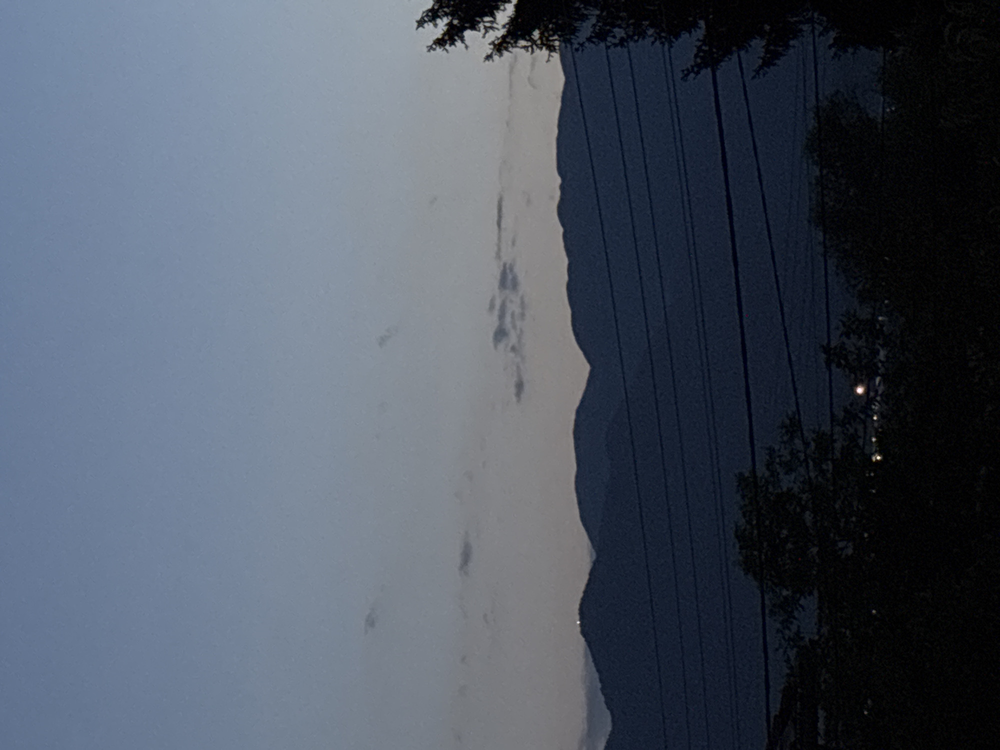 The image depicts a serene twilight or nighttime landscape, likely in Garmen, Bulgaria, given the provided context. The sky occupies the upper portion of the frame, displaying a gradient from a lighter, muted blue-grey at the top to a slightly darker tone towards the horizon. A few scattered, faint clouds are visible against the sky, suggesting a partly cloudy evening.
A prominent mountain range forms a dark, undulating silhouette across the lower middle of the image, sharply contrasted against the lighter sky. The peaks and slopes of the mountains are distinct despite their darkness. In the foreground, particularly towards the right side, the dark outlines of dense trees and foliage are visible, indicating a forested area. Several parallel power lines stretch across the lower right section of the image, running diagonally and partially obscuring the view. A few small, bright points of light, possibly distant artificial lights from a settlement or infrastructure, can be discerned within the dark mass of the mountains or foliage in the lower right.
No specific activity or people are visible in the scene, which conveys a sense of quietude and stillness. The time of day is clearly dusk or night, characterized by the low light and silhouetted features. There is no visible text in the image. The overall impression is one of natural beauty and tranquility in a mountainous, rural setting.