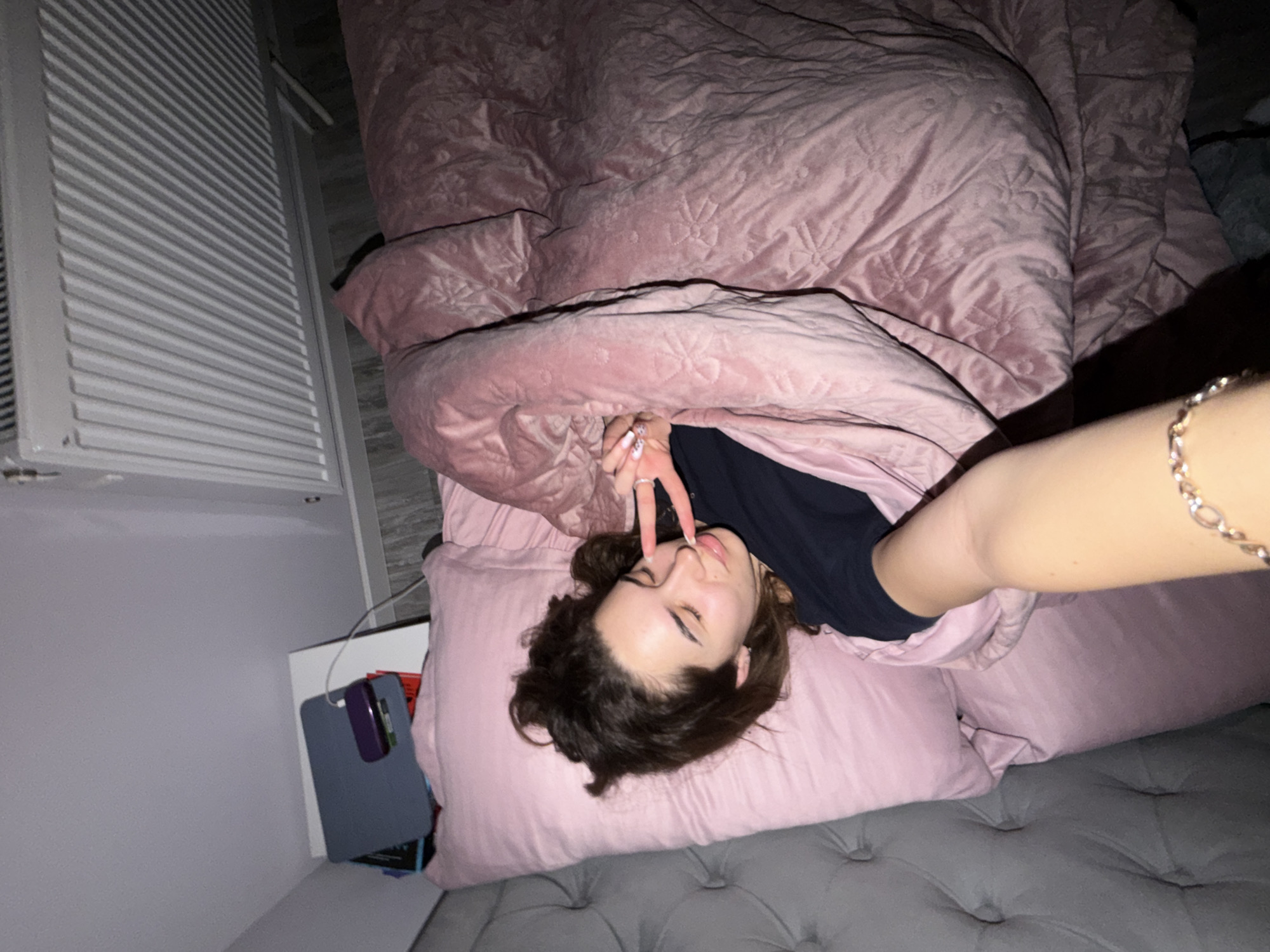 A young woman is lying on a bed under a pink quilt, holding two fingers to her lips in a "V" gesture. She has dark hair and is wearing a dark shirt. The bed has a pink pillow and is placed on a grey button-tufted mattress. A grey radiator and a small table with books or other items are visible in the background. The location in Dobrich, Bulgaria cannot be determined from this image.