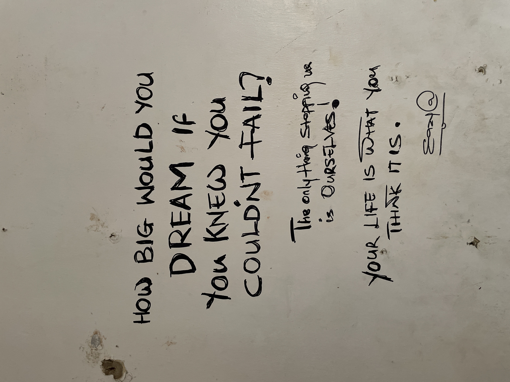 The image displays a white, textured wall with black marker graffiti. The graffiti consists of three distinct phrases and a signature. The first phrase, written in large, bold letters, asks, "HOW BIG WOULD YOU DREAM IF YOU KNEW YOU COULDN'T FAIL?". Below this, a smaller, cursive phrase reads, "The only thing stopping us is OURSELVES!". The third phrase, also in cursive, states, "YOUR LIFE IS WHAT YOU THINK IT IS.". The signature at the bottom appears to be "Eazy@". There are several small holes and marks on the wall, consistent with age or damage. The lighting suggests an indoor setting with a diffused light source. There are no people or animals visible in the image. The context provided about Ado, Nigeria, is not visually supported by the image itself.