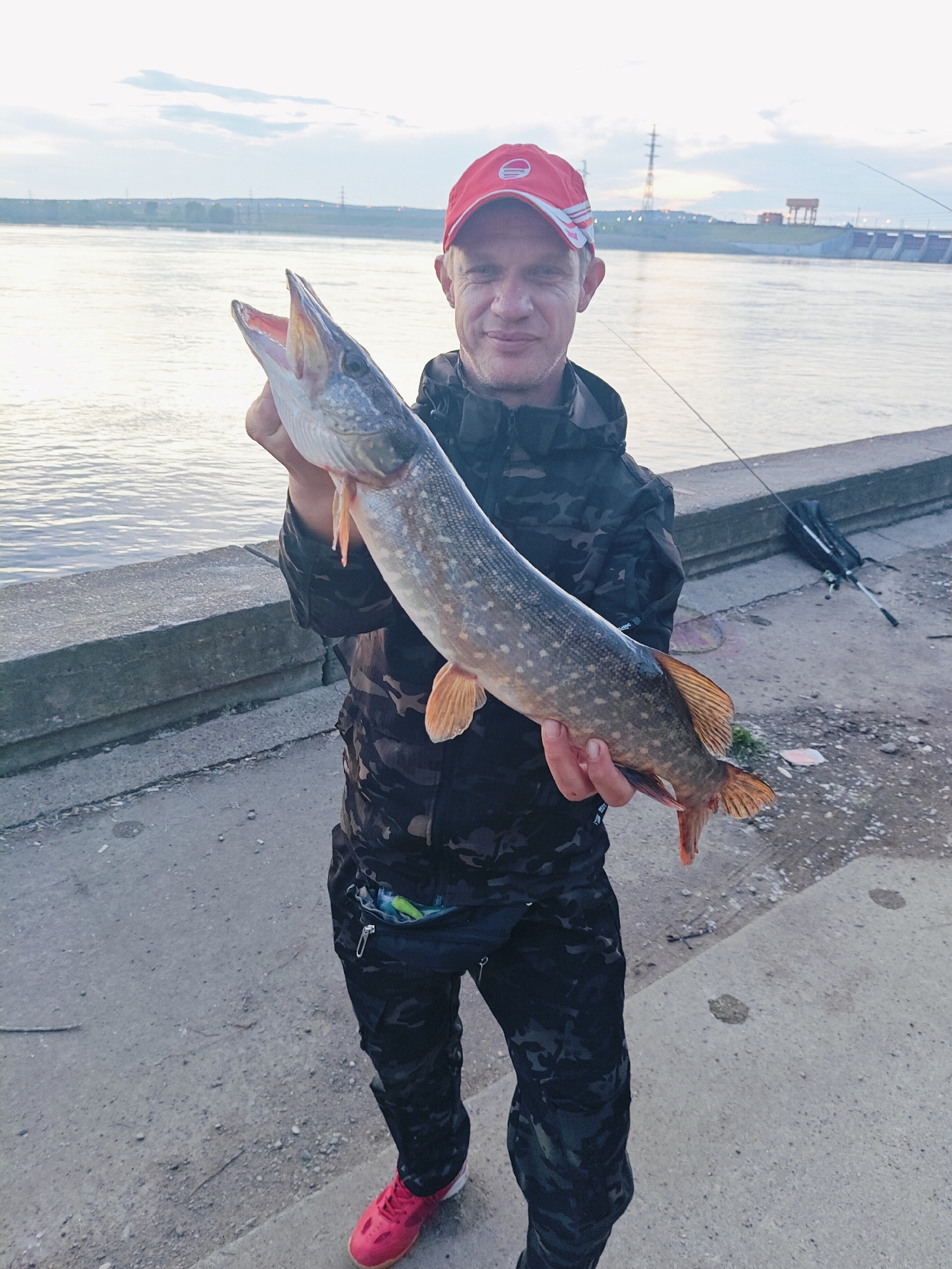 A man wearing a red and white baseball cap and a camouflage jacket and pants is holding a large pike fish by the river. He is standing on a concrete embankment. In the background, there is a river, a dam, and a few buildings and power lines. Two fishing rods are leaning against the embankment behind him. The sky is cloudy. The image appears to have been taken in the city of Chaikovsky, Russia, near the Kama River and the Votkinsk Dam.