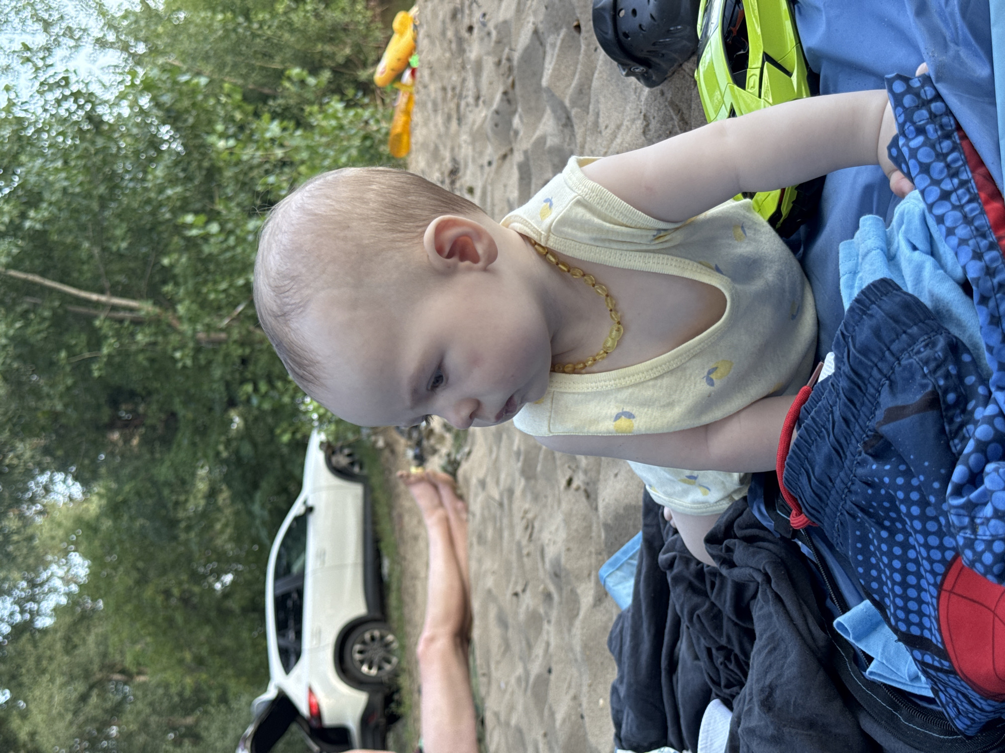 A young child, wearing a yellow sleeveless shirt and a necklace, lies on a blue blanket on a sandy surface. Behind the child, a white SUV with its trunk open is parked on a grassy area bordering trees. The child is facing to the left, with their body angled towards the viewer. The surrounding environment appears to be a park or recreational area, possibly a beach, given the presence of sand and water elements in the background. There are various clothing items and a yellow toy airplane scattered on the sand.