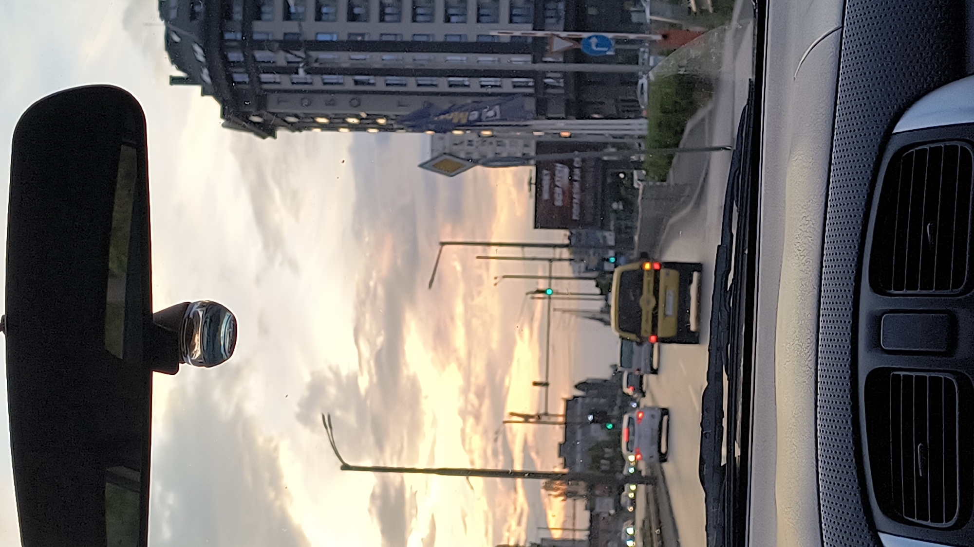 The image, taken from inside a vehicle, captures a view of a street in Sofia, Bulgaria at sunset. The orange and pink hues of the setting sun are reflected in the sky. In the foreground, a side mirror and a dashboard element frame the view. Through the windshield, a street is visible with multiple vehicles, including a yellow van, waiting at what appears to be a green traffic light. Buildings line the street, with commercial signs. There are street lamps and traffic signals along the road.
