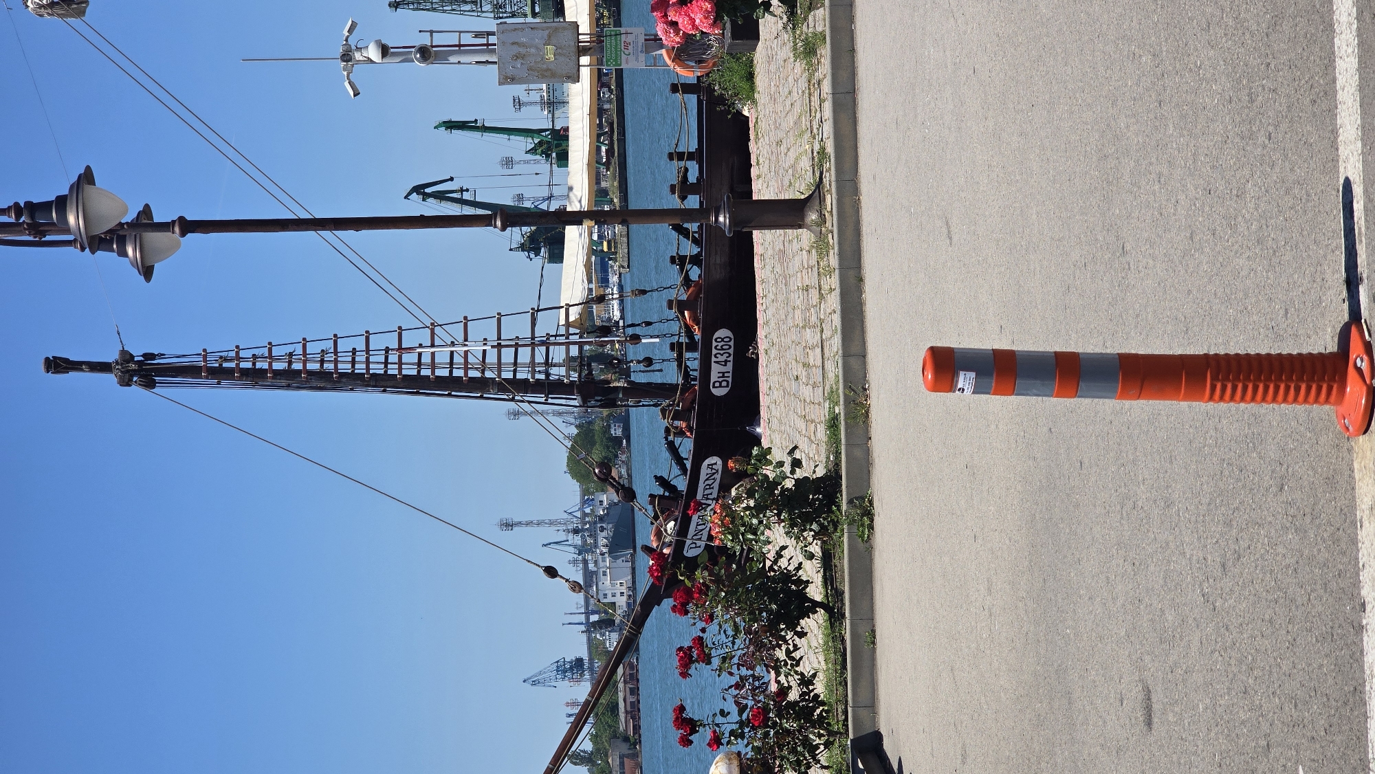 The image shows a view of the Varna port area in Bulgaria. In the foreground, there is a red and grey traffic bollard on an asphalt surface. Behind the bollard, a cobblestone promenade runs along the waterfront. A wooden ship with a tall mast and rigging is docked at the promenade. The ship has the name "PANTARNA" and the registration "BH 4368" visible on its side. Red roses are blooming along the edge of the promenade. In the background, the blue waters of the port are visible, with various cargo cranes, industrial structures, and other ships present. The sky is clear and blue, indicating a sunny day. The overall scene depicts a bustling port environment with elements of maritime history.