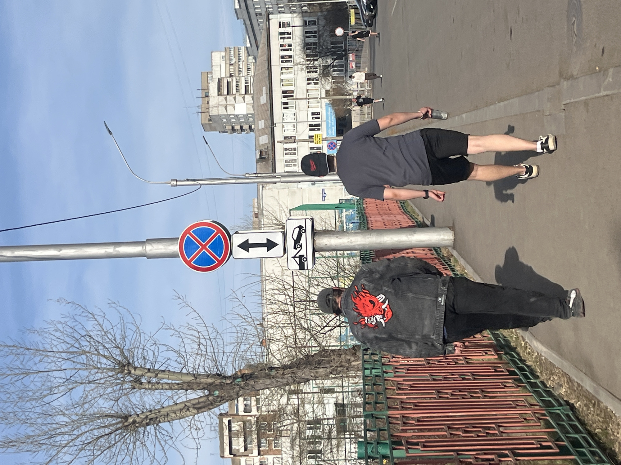 The image shows an outdoor scene in Krasnoyarsk, Russia, during daylight hours. Two male figures are walking along a sidewalk adjacent to a street. The first figure, closer to the camera, wears a black hat and a black denim jacket with a large red and white design on the back. The second figure wears a black baseball cap and a gray t-shirt, and black shorts. Buildings line the opposite side of the street. A traffic signpost with a "no parking" symbol and directional arrows indicating opposite directions stands near the sidewalk. A metal fence runs along the edge of the sidewalk in the foreground. A bare tree is also present. The sky is blue with minimal cloud cover. Other pedestrians and vehicles can be seen farther down the street.