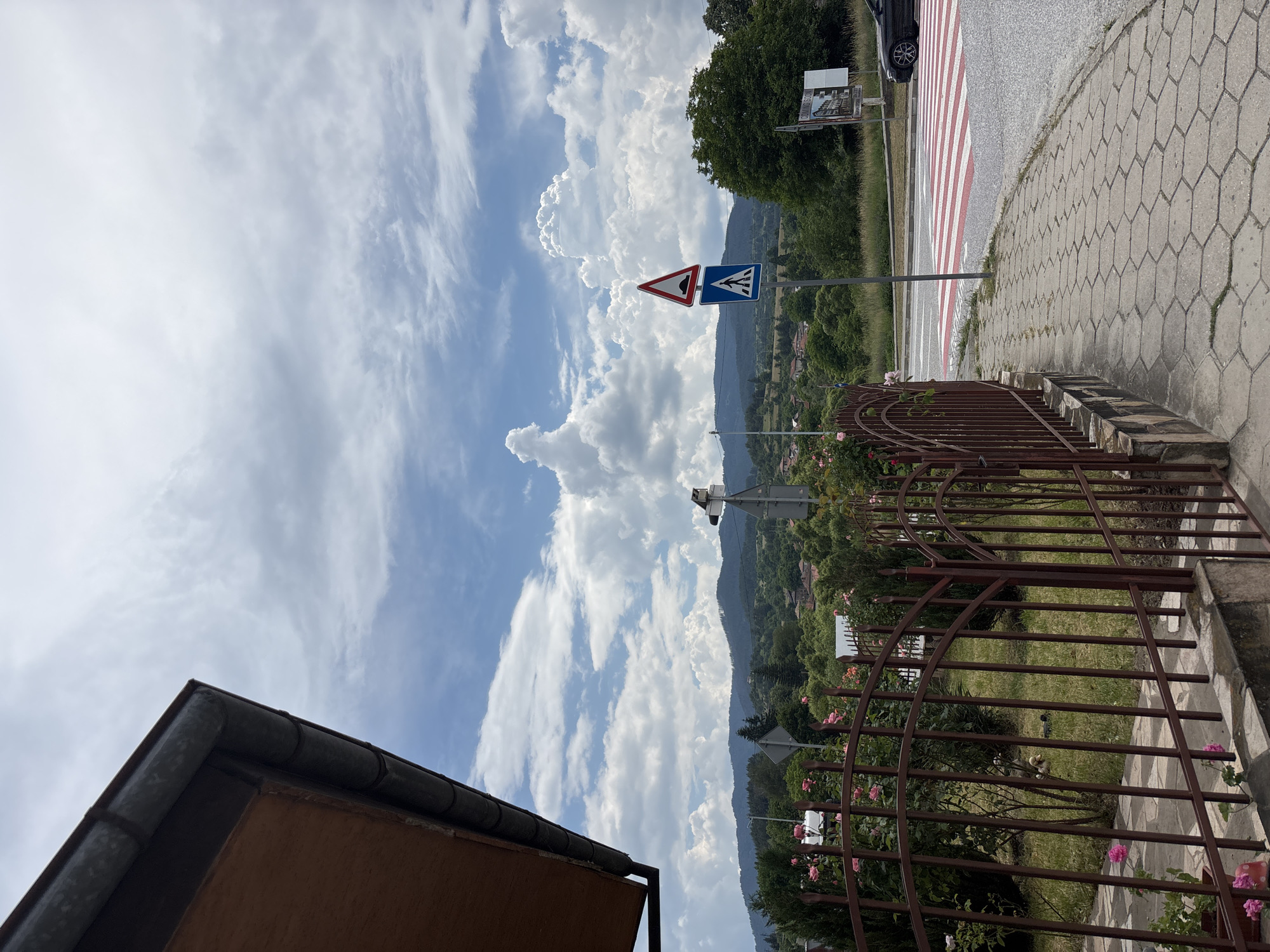The image depicts a street scene in Hvoina, Bulgaria, under a partly cloudy sky. In the foreground, a brown metal fence lines a paved sidewalk with hexagonal concrete tiles. Beyond the fence, a grassy area with pink roses and green bushes is visible. A red and white striped crosswalk stretches across the road in the mid-ground, with a dark-colored car stopped on the right side of the crosswalk. Traffic signs indicating a speed bump and a pedestrian crossing are mounted on a pole. In the background, rolling green hills dotted with houses and trees create a scenic landscape. The sky is filled with dramatic cumulus clouds, some of which are illuminated by sunlight.