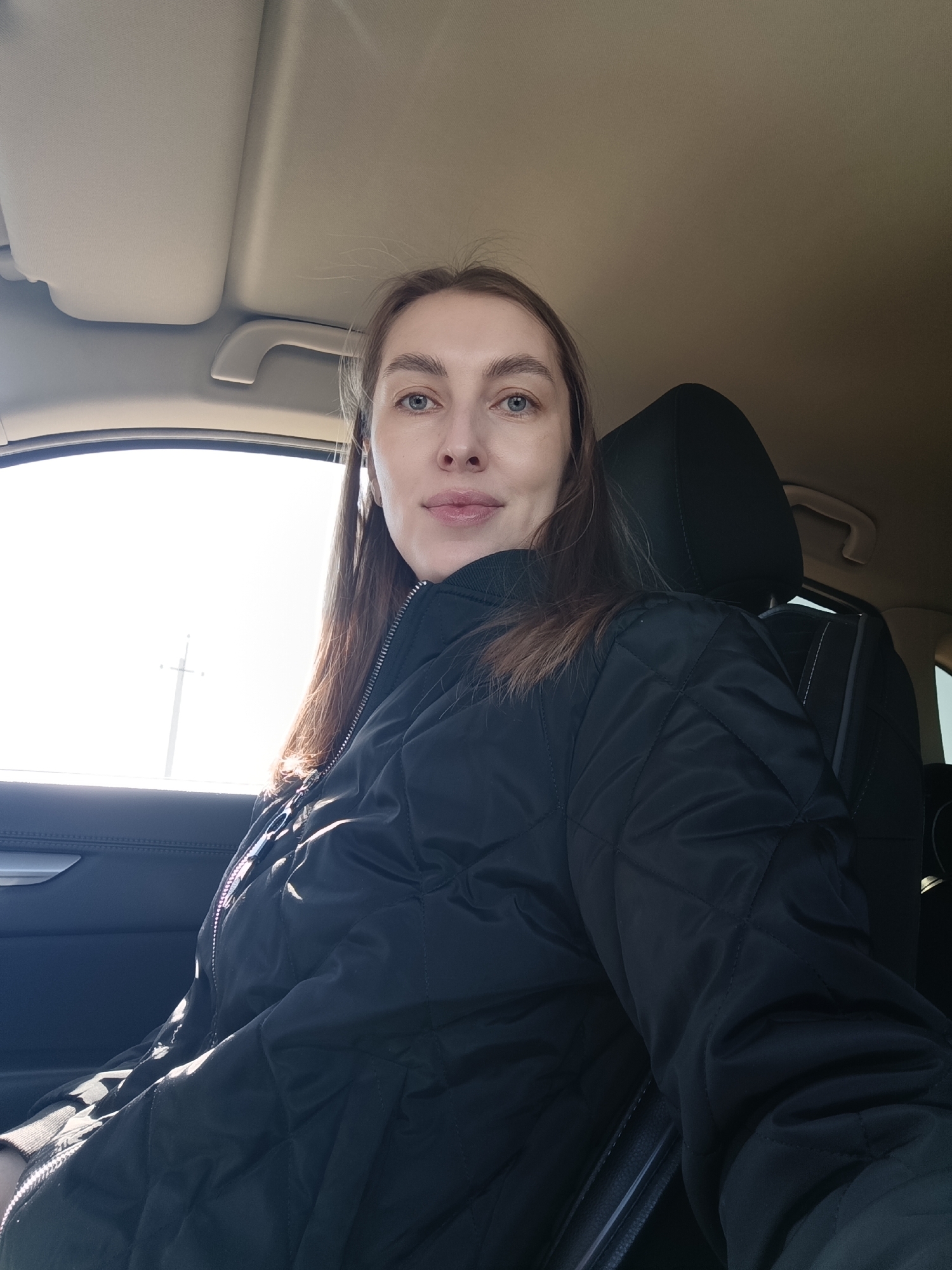 A woman is sitting in the driver's seat of a car. She is wearing a black quilted jacket. The interior of the car is dark, with a light-colored roof and dashboard. The window is open and shows a bright, sunny day. Outside the window there is a white light post. Based on the location of Chaikovsky, Russia, it may be implied that the woman is Russian.