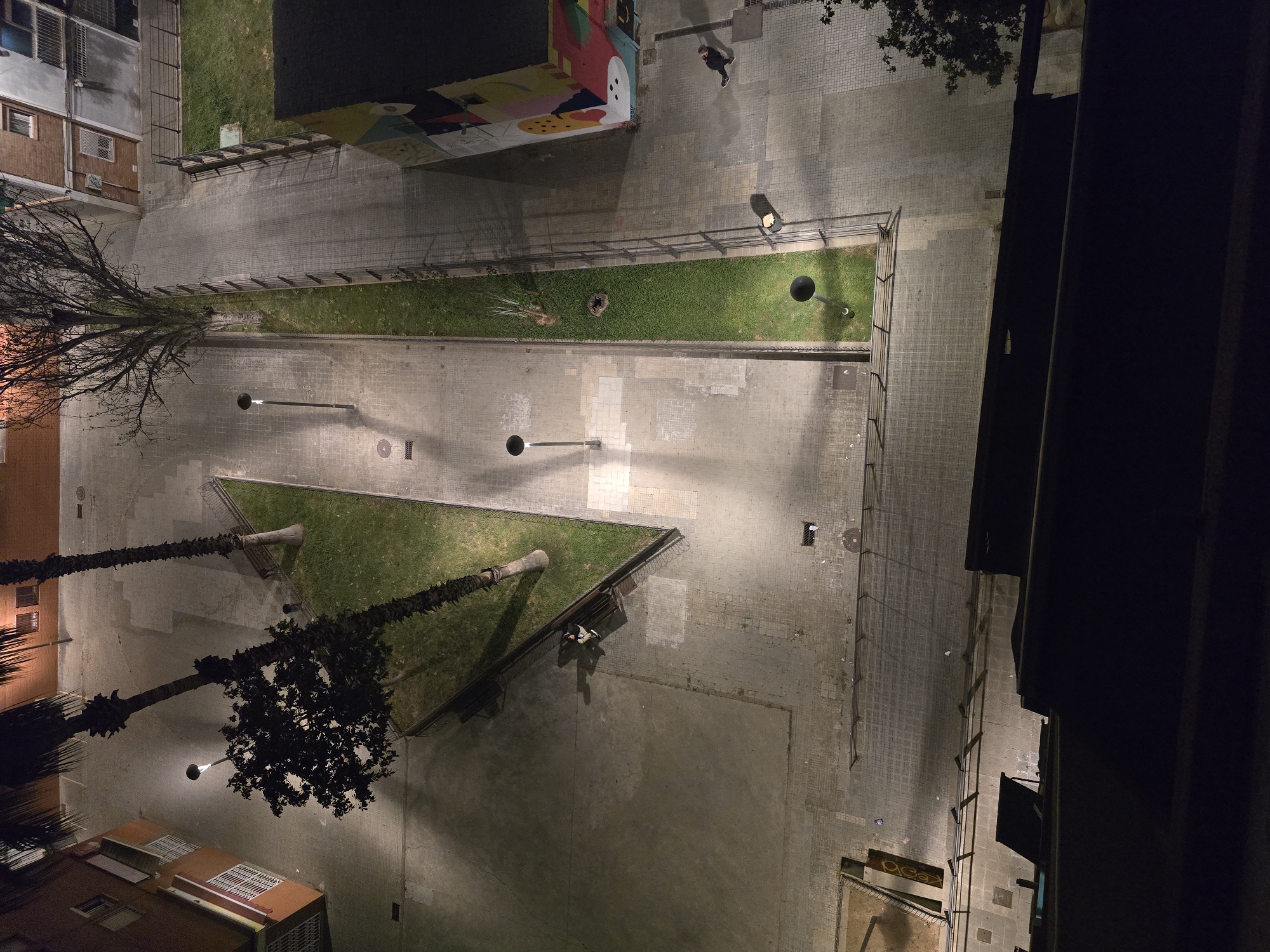 An aerial view shows a streetscape in Cornellà de Llobregat, Spain, at night. The scene features a T-junction of paved walkways, sectioned with linear green spaces. A mural adorns a wall at the top of the image. Streetlights illuminate the area. Trees are planted, some bare. At the very top a pedestrian is walking. Several benches are seen. Building facades are visible on the sides of the image.