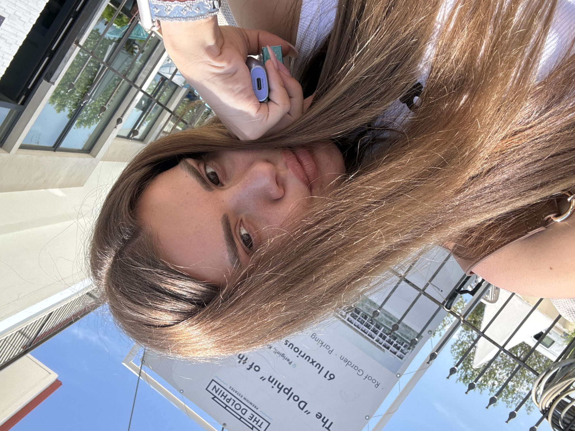 A woman is shown in a self-portrait, holding a small electronic device. The background features a banner for "The Dolphin Premium Estates" with text reading "The 'Dolphin' of th" and mentioning "61 luxurious Roof Garden Parking" in Perigiali, Unknown. A building with glass windows is visible behind her, and the sky is blue.