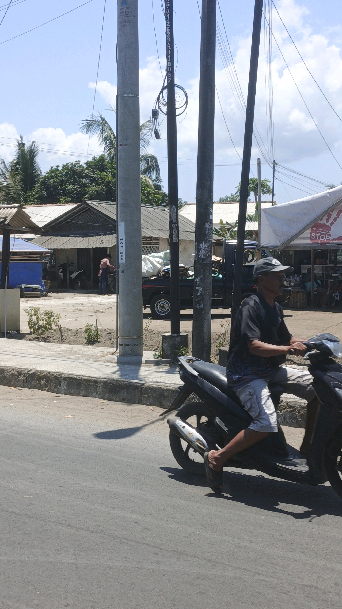 A man wearing a dark t-shirt, patterned shorts, and a baseball cap rides a black motorcycle on a paved road. He is looking to his right. The motorcycle has a silver exhaust pipe and a black seat. In the background, there are several utility poles, trees, and buildings with corrugated roofs. One building has a white awning with the word "STOP" written in red. There is also a black pickup truck in the background. The scene appears to be in a sunny, outdoor urban or semi-urban environment, potentially in Pangandaran, Indonesia, given the architecture and vegetation.