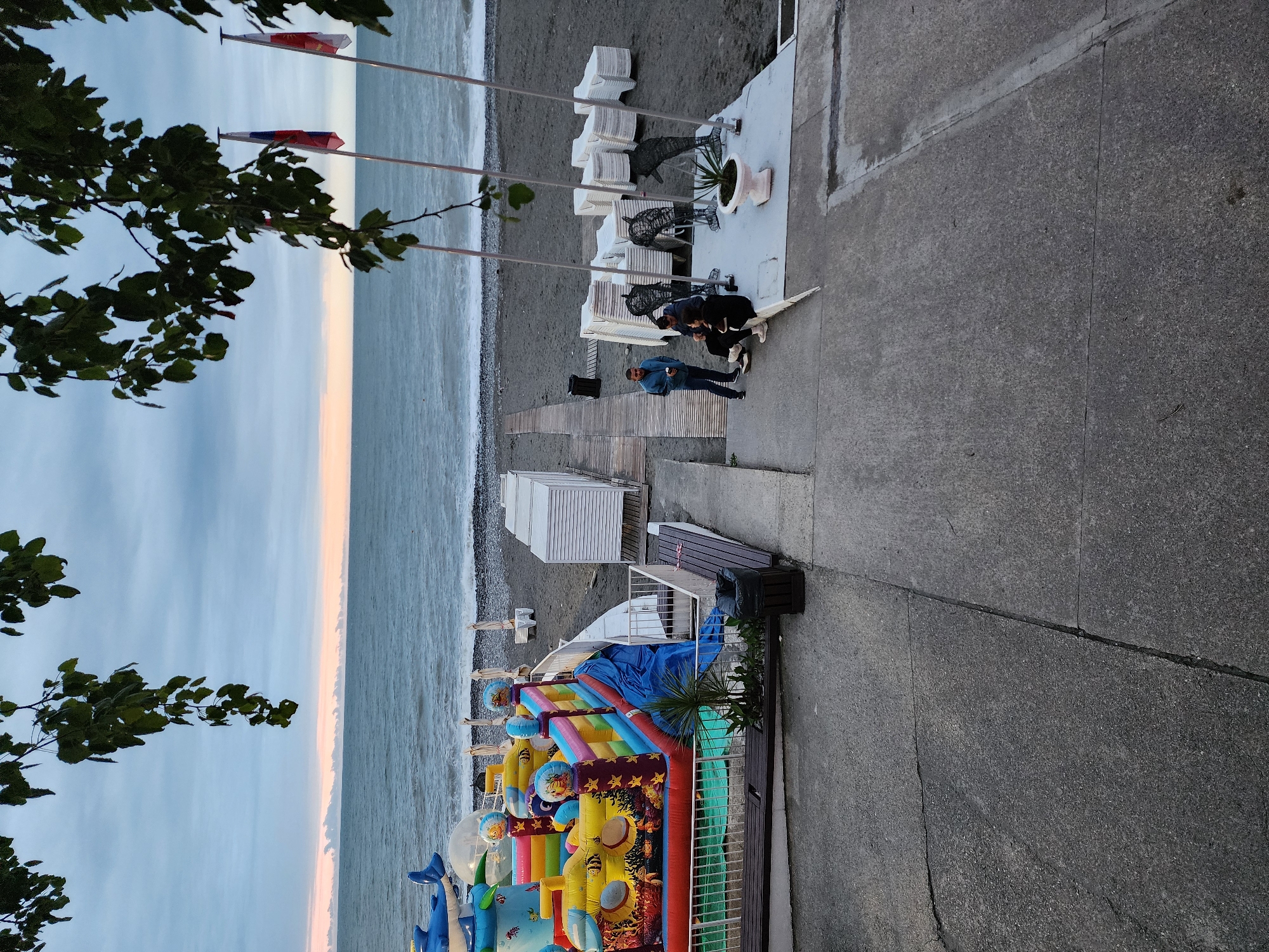 The image depicts a beach scene in Sochi, Russia, angled vertically. The Black Sea meets a gray, pebbly beach. Three flagpoles stand along the beach. A paved area runs parallel to the beach, featuring stacked white lounge chairs and several individuals. One man in a blue jacket stands observing two people seated on a bench. An inflatable, colorful playground structure sits closer to the foreground of the image. The sky is overcast, with a faint hint of pink on the horizon. Trees frame the left side of the image.