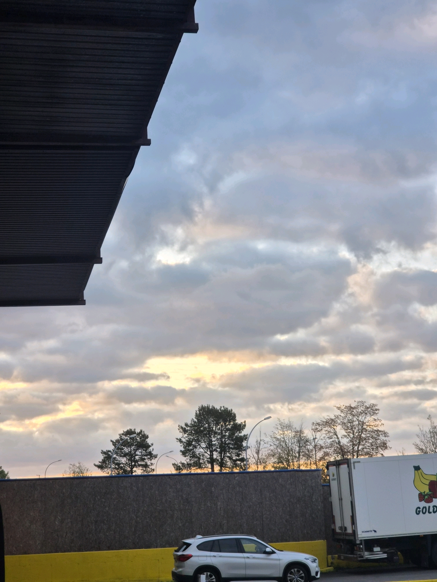 The image depicts an outdoor scene, likely in a semi-industrial or commercial area. Dominating the upper left is the corrugated metal underside of a structure, possibly a warehouse or covered parking. The sky above is filled with a mix of gray and white clouds, with hints of orange and yellow suggesting either sunrise or sunset.
In the mid-ground, a dark wooden fence runs horizontally across the frame, partially obscured by trees. Several streetlights are visible behind the fence, with one prominent one arching over a dark green pine tree. Further to the right, the top portion of a white refrigerated truck is visible, featuring a logo with cartoonish depictions of bananas and strawberries, along with the partial word "GOLDEN". Below the fence, a bright yellow barrier runs along the ground.
In the foreground, a silver SUV is parked. The overall impression is of a commercial or logistical hub under a dramatic sky. Based on the visual elements, the location is likely Leudelange, Luxembourg, given the characteristic architectural styles and vegetation often found in such areas of the country. No people are present in the image.