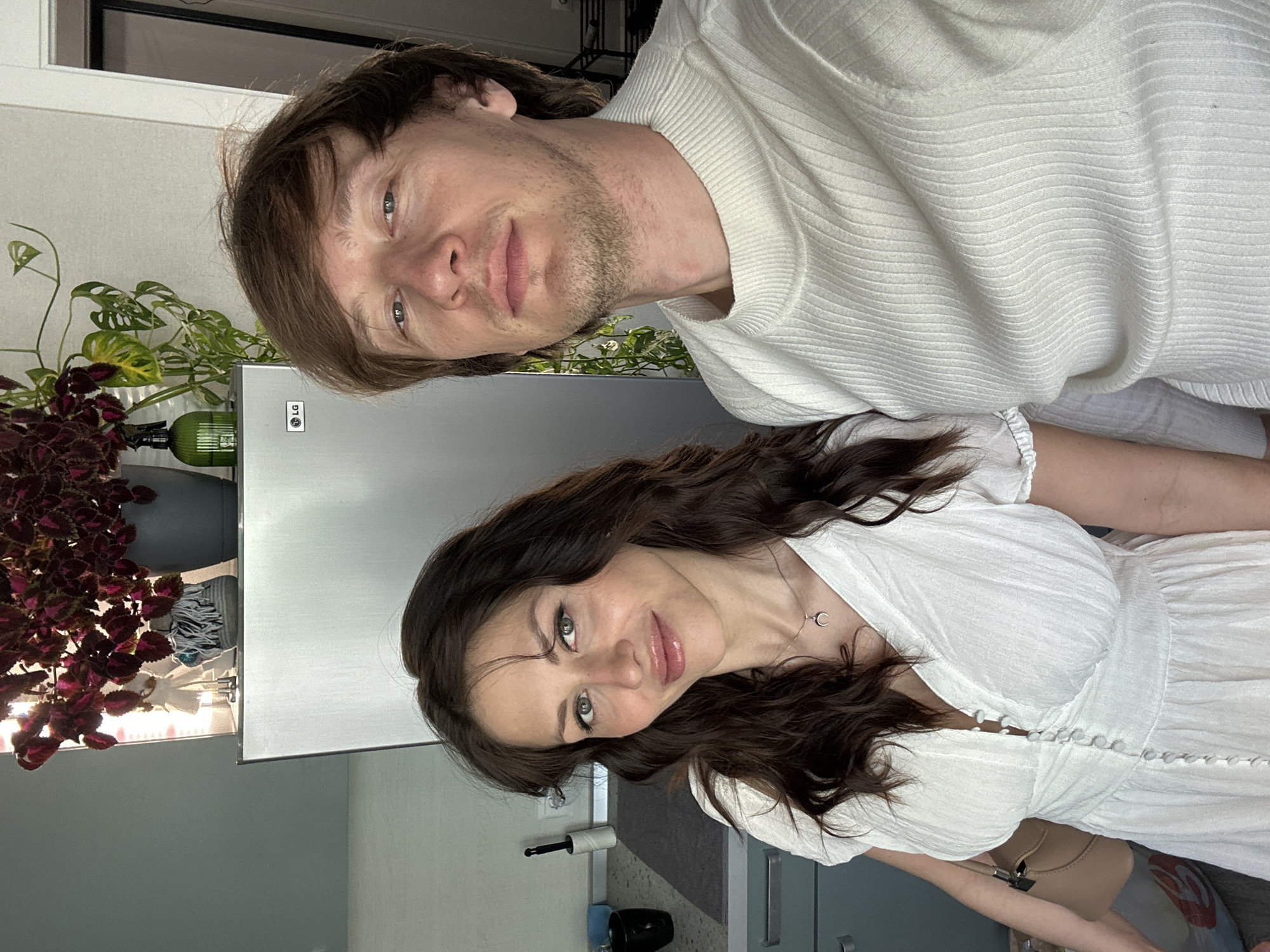 Two individuals, a male and a female, are posing indoors. The man, positioned at the top of the frame, is fair-skinned with brown hair and a light beard. He is wearing a white sweater. The woman, positioned at the bottom, has long brown hair and is wearing a white dress.
Behind them is a silver LG refrigerator. To the left of the refrigerator, there are potted plants. The surroundings suggest an interior setting, likely a kitchen or living area. The location is not evident.