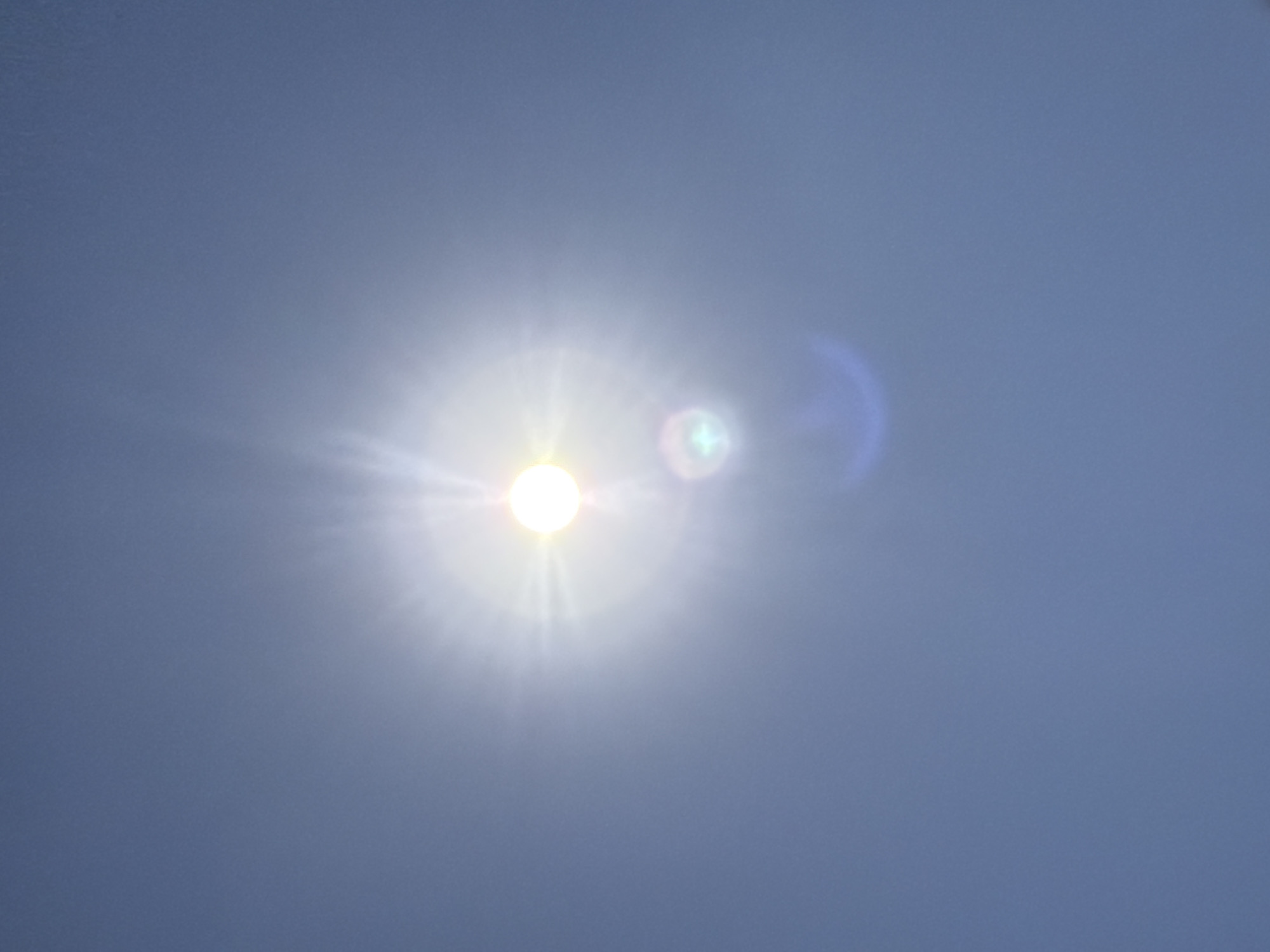 The image is a close-up, vertical shot of the sun and sky. The sun is the brightest object in the frame, emitting intense light rays and causing lens flare. There are also some circular lens flares, one of which has a greenish hue, and a blueish arc of light. The sky is a clear, pale blue. The image appears to be taken outdoors during the day, likely with the camera pointed upwards. There are no visible structures or any indication of a city, including Sharm El Sheikh, Egypt.