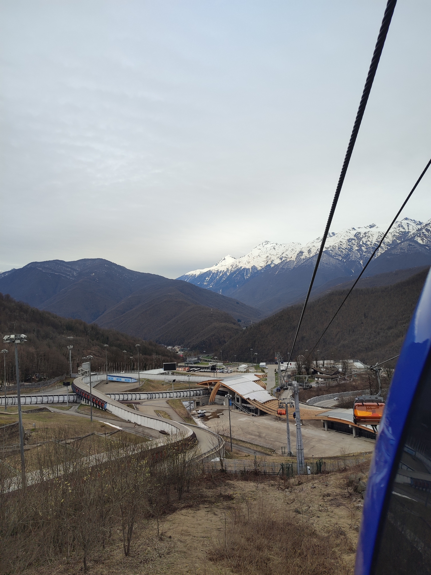 The image captures a view from a ski lift in город Сочи, Russia. The scene is dominated by a bobsled track winding through a valley, with the track structure painted with blue and red accents. Several support structures are visible around the track. A modern building with a light-colored, angular roof is situated near the track, appearing to be a station or facility related to the bobsled run. Ski lifts are operating in the foreground and mid-ground, with cabins of orange and blue colors visible. The background features a mountain range with snow-capped peaks. The sky is overcast, casting a diffuse light over the landscape. The lower portion of the image contains vegetation in muted brown tones, suggesting either a transition from summer to autumn or a dormant state due to winter conditions. The overall impression is of a mountainous landscape designed for winter sports, captured from a high vantage point.