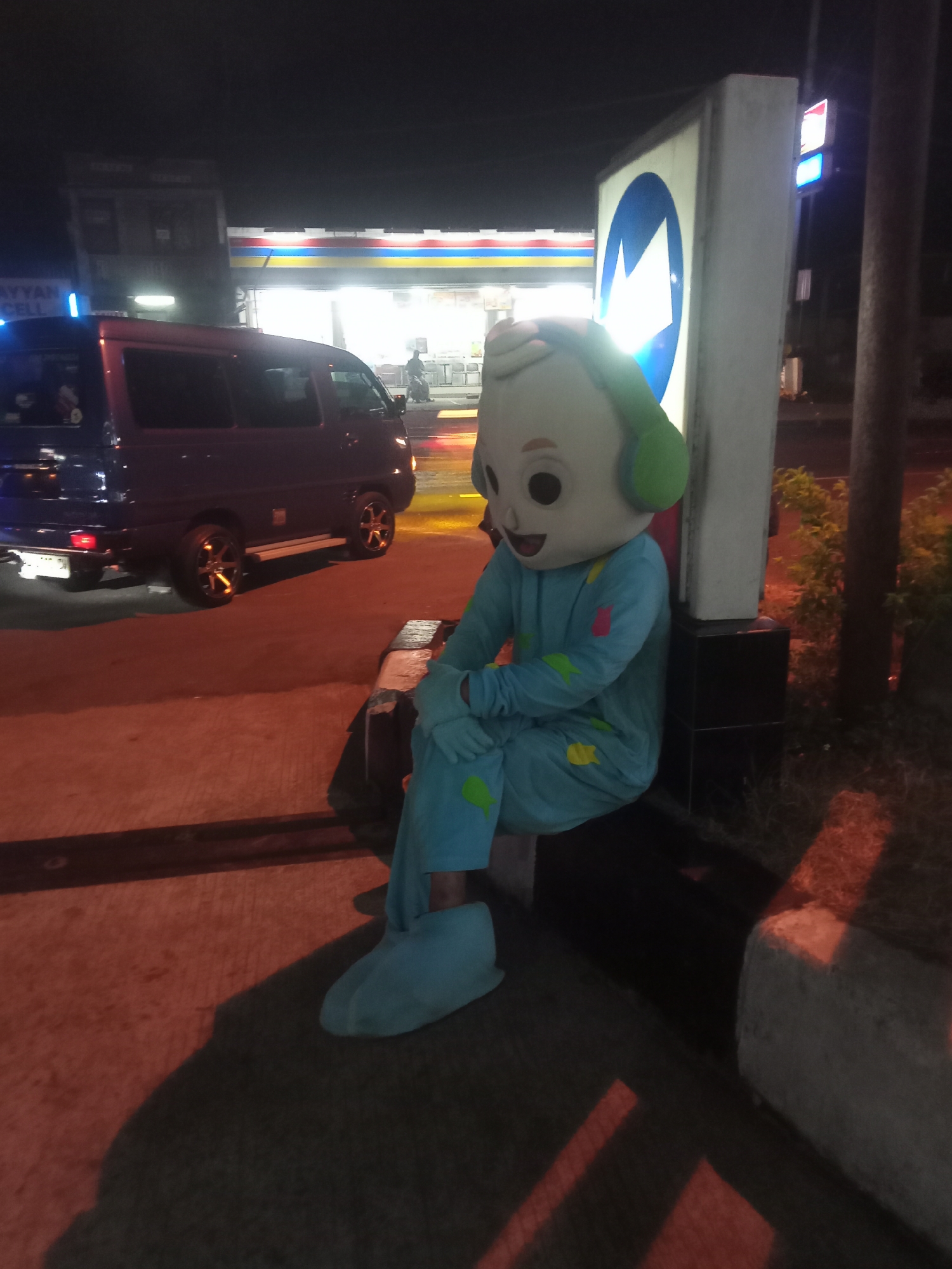 A person wearing a light blue cartoon character costume with green headphones sits on a concrete bench at night. The costume features a large head with a smiling face, light blue eyes, and a light green mouth. The suit has yellow and pink shapes on the sleeves and legs. The character is leaning against a tall, white, rectangular sign with a blue circle and a white arrow pointing to the right. A dark purple van is parked to the left of the frame. In the background, a brightly lit convenience store is visible, with a yellow, red, and blue striped awning above the entrance. The scene appears to be a street in Gladagsari, Indonesia, at night, with streetlights and the store's illumination providing light.