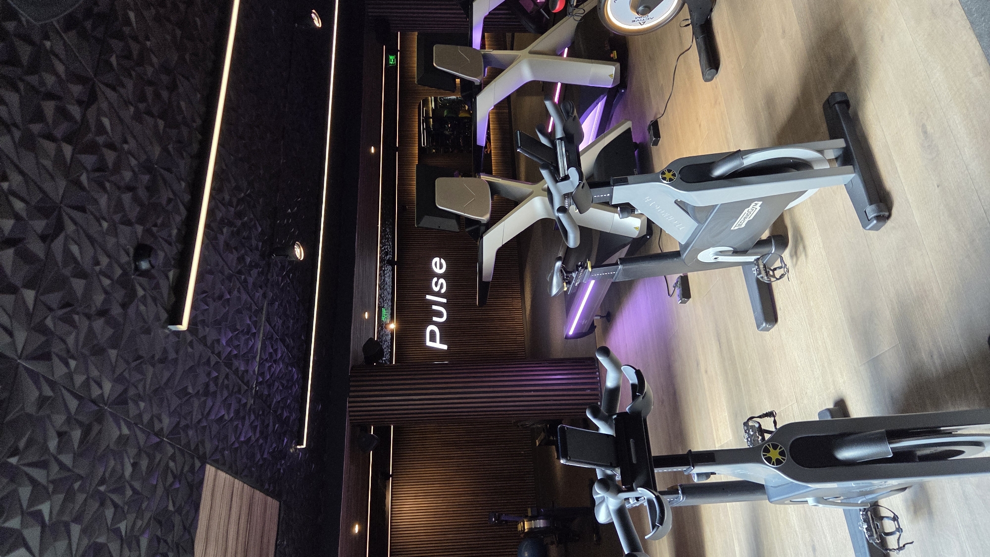 The image shows the interior of a fitness center or gym in Unknown, Unknown. The scene is oriented vertically. Three stationary bikes are visible on a light brown wooden floor. The bikes are primarily gray and black, with some white accents. They are arranged in a line, suggesting a possible workout class setup. The "Pulse" logo is prominently displayed on a wall behind the bikes. The background wall features a textured, geometric pattern in a dark color and horizontal striped wooden panels. A line of lights runs along the wall. Purple lighting accents the area. The focus is on the bikes and the gym's interior design, with no people visible.