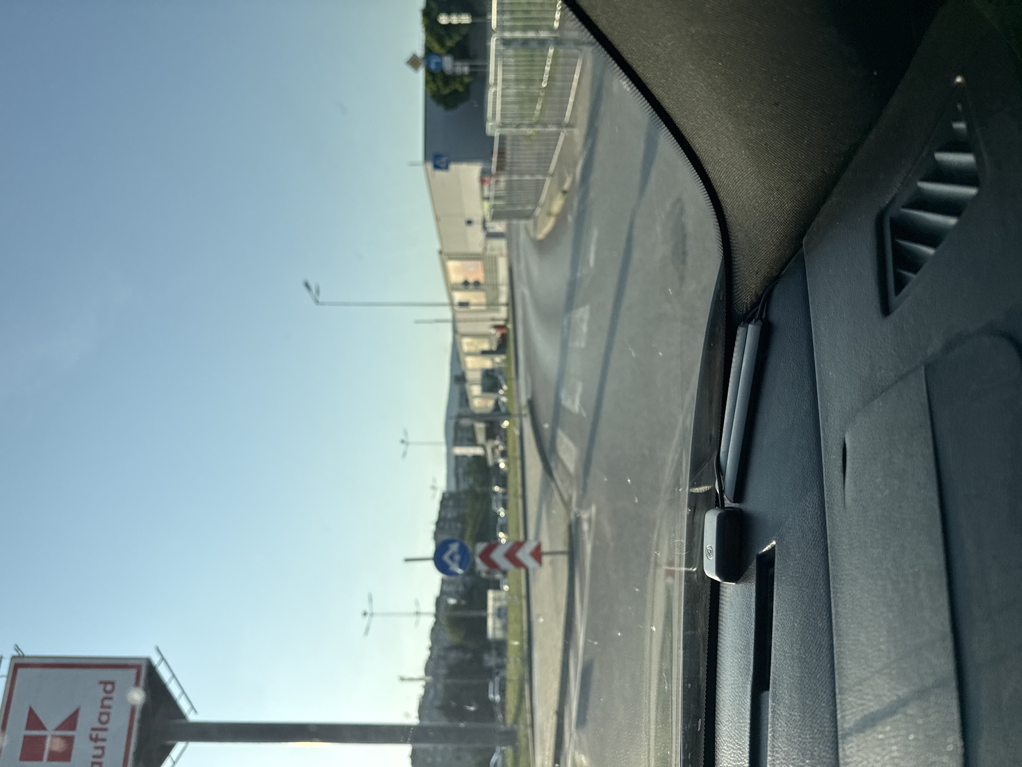 The image captures an outdoor, urban scene from the perspective of a vehicle's dashboard, likely approaching an intersection or a commercial entrance. The setting is Stara Zagora, Bulgaria, specifically near the Kaufland store located at bul. Nikola Petkov 48.
In the foreground, the dark grey, textured dashboard of a vehicle is visible, occupying the bottom third of the frame. A small, black rectangular device, possibly a GPS unit or part of a dashcam, is positioned on the dashboard with a dark cable extending from it. Adjacent to it lies a slender, dark object, possibly a pen. An air vent with vertical slats is visible on the far right of the dashboard. The top edge of the windshield shows the vehicle's interior trim.
The midground features an asphalt road with white markings, including a clear pedestrian crossing. The road curves slightly to the right. A prominent red and white chevron sign, indicating a sharp curve or obstacle to the right, is positioned on a central island. Above and slightly to the left of the chevron sign, a blue circular sign with two white arrows (one pointing upward and curving right, the other downward and curving left) denotes a mandatory direction or passage. Further along the road on the right, silver metal barriers separate the roadway from what appears to be a sidewalk or parking area. A blue rectangular sign with a white pedestrian symbol indicates a pedestrian crossing.
In the background, a large, light-grey commercial building, identifiable as a Kaufland supermarket by its distinctive red "K" logo and partial "aufland" text on a tall sign, dominates the left side. Other commercial buildings are visible further down the street on the right. In the far distance, residential buildings and gentle hills or mountains are silhouetted against the horizon. Tall streetlights with multiple arms are scattered throughout the scene. The sky is clear and light blue, suggesting good weather, and the lighting indicates either morning or late afternoon sun, casting soft shadows. There are no clearly visible individuals in the scene.