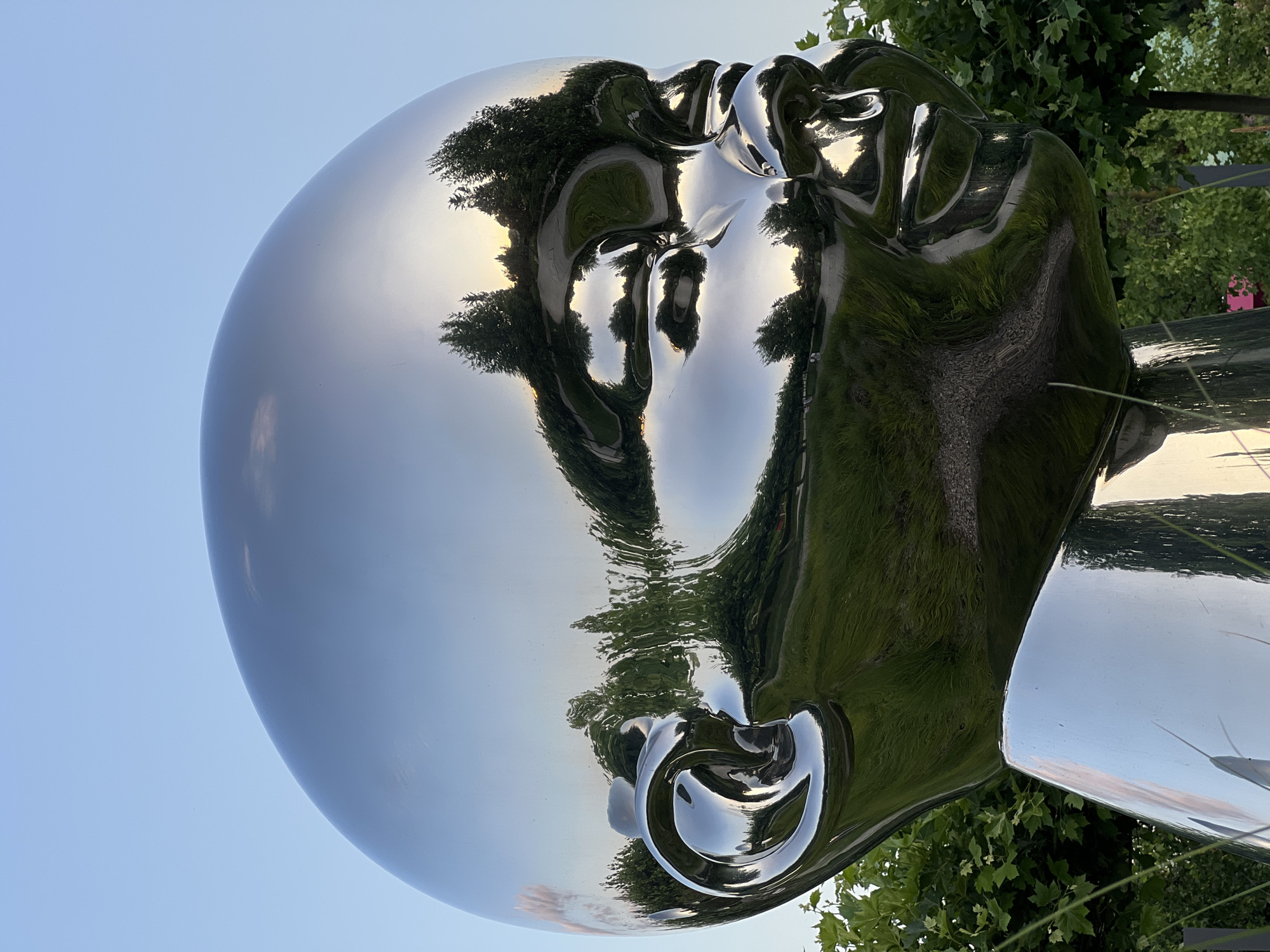 The image features a highly polished, abstract sculpture of a human head, rendered in a reflective metallic material. The sculpture is positioned outdoors, with lush green trees and a clear sky visible in its distorted reflections. The light suggests it is daytime, likely late afternoon or early evening due to the soft, warm tones of the sky. The sculpture's surface acts like a mirror, reflecting the surrounding environment, including trees with dappled sunlight filtering through their leaves, and a pathway or road. The facial features are stylized and smooth, with the curves of the head creating a sense of gentle motion or flow. The background hints at a park or garden setting, with the presence of foliage and what appears to be a clear sky. The overall mood is serene and contemplative, amplified by the smooth, almost liquid reflections. There is no visible text in the image.