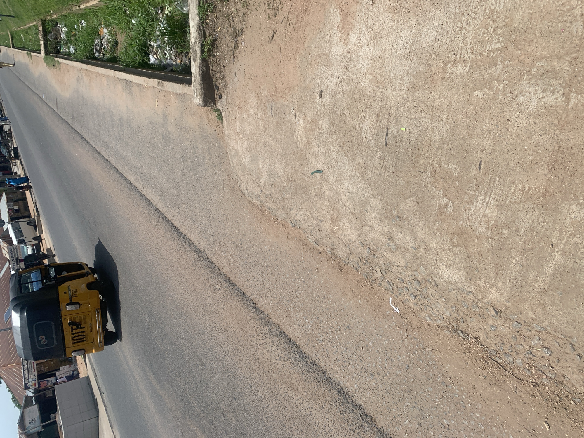 This media file shows a street scene in Jalingo, Nigeria. A yellow and black auto-rickshaw, also known as an "okada" or "keke napep," is driving down the road. The rickshaw has the number "101T" printed on its side in large, white numerals, along with some other text that is difficult to read. The road appears to be paved and is lined with buildings and businesses on the left side of the frame. On the right side of the road, there is a raised concrete curb and an area of unpaved ground with some scattered debris. In the distance, there are more buildings and a glimpse of greenery. The lighting suggests it is daytime, likely with clear weather. There are no people clearly visible in the frame besides the driver of the rickshaw. The overall impression is of a typical street scene in a Nigerian city.