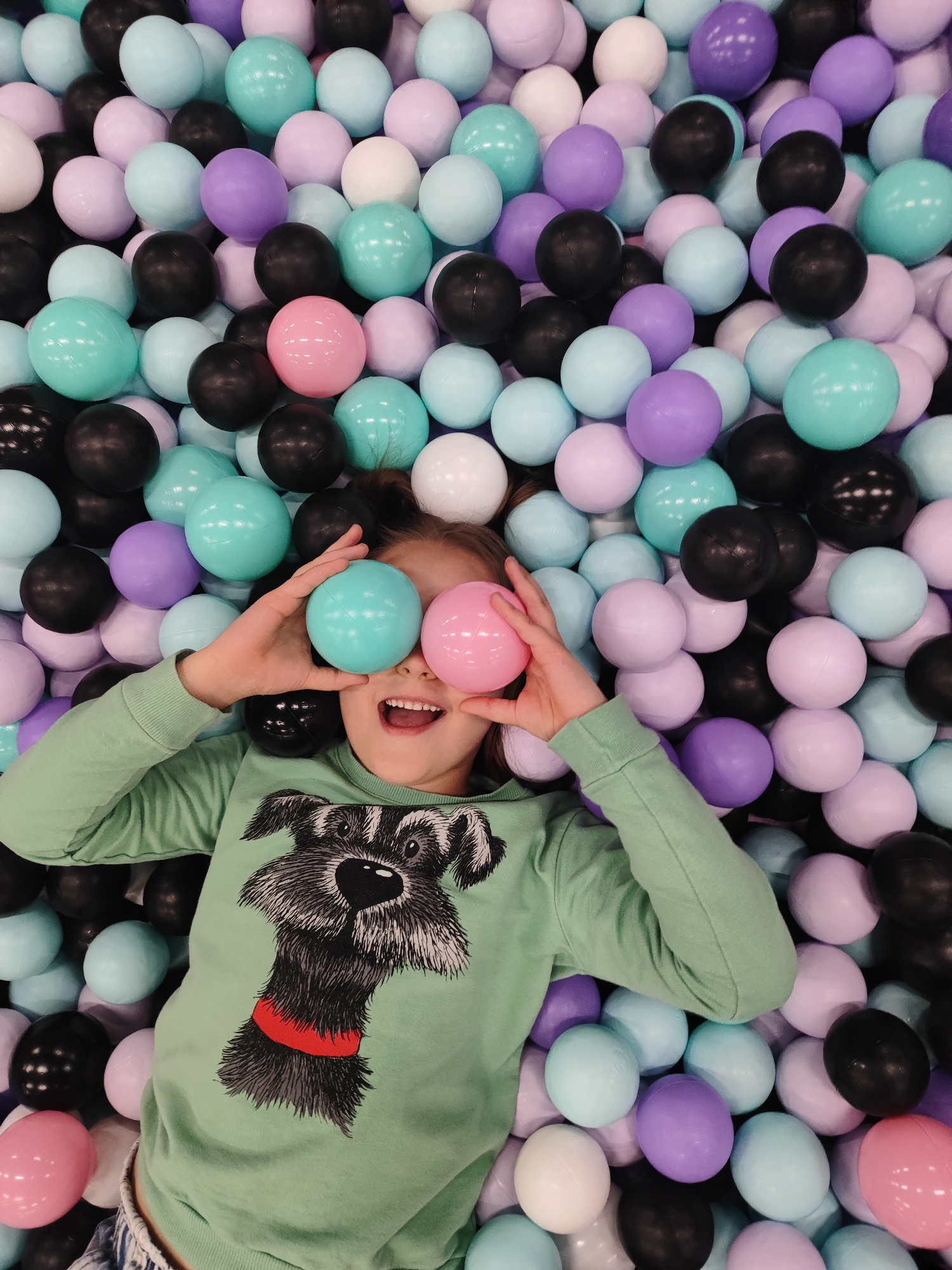 A young girl lies supine in a ball pit, holding a pink and a blue ball in front of her eyes. She is wearing a light green long-sleeved shirt with a black and gray dog image. The ball pit contains black, light blue, lavender, pink, and white balls. The scene is brightly lit. The girl is smiling and appears to be enjoying herself. No specific location in Krasnodar, Russia can be determined from the image.