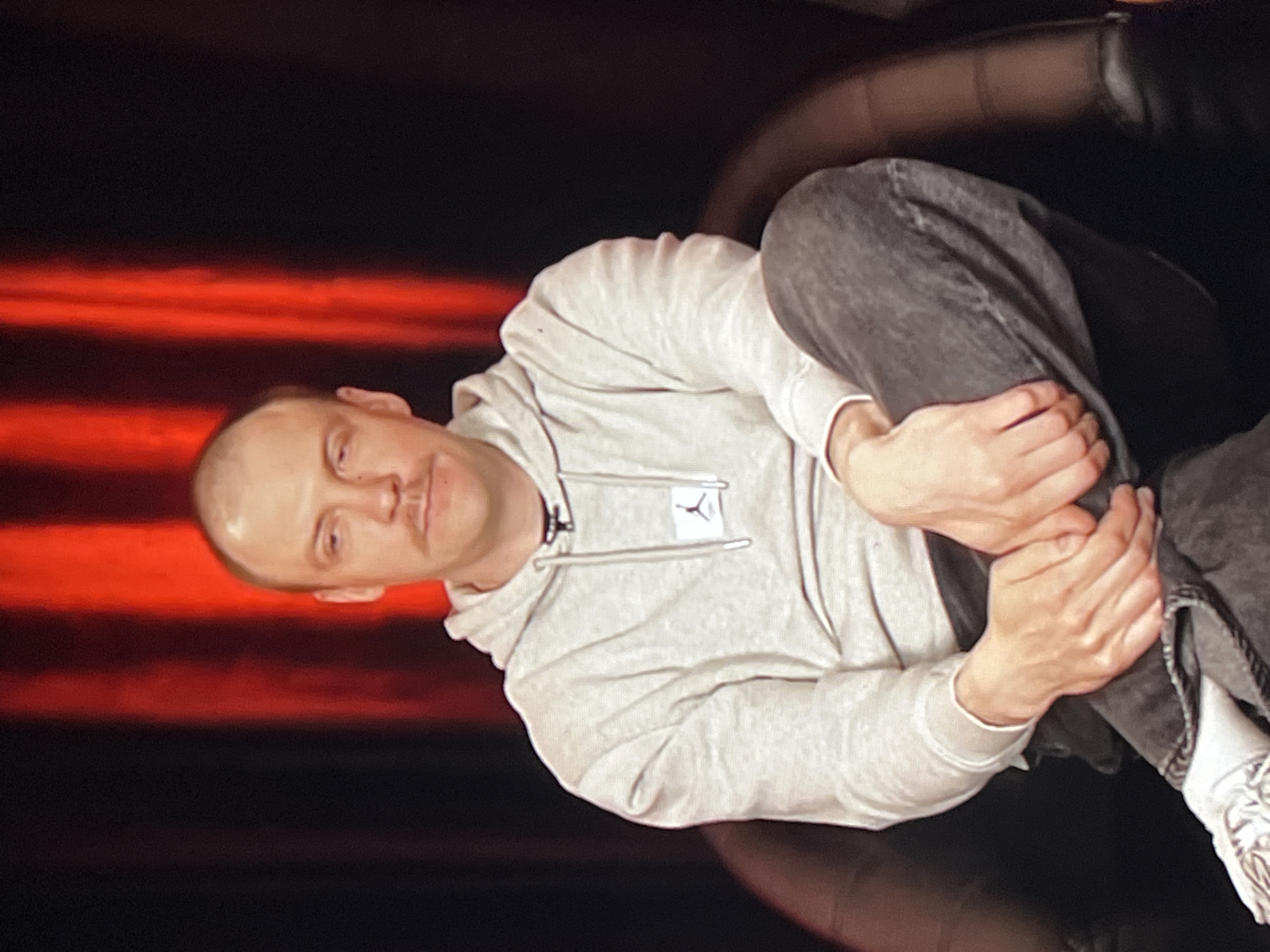 A medium shot captures a bald man with a mustache sitting in a dark chair. He is wearing a light grey hoodie with a small black logo on the left chest and dark grey jeans. His left leg is crossed over his right, with his hands resting on his lap. He is looking slightly to his left with a neutral expression. The background consists of dark curtains with vertical red lighting. The image appears to be a still from a video recording or photograph taken in an indoor studio setting, possibly in Genemuiden, Netherlands, given the context.