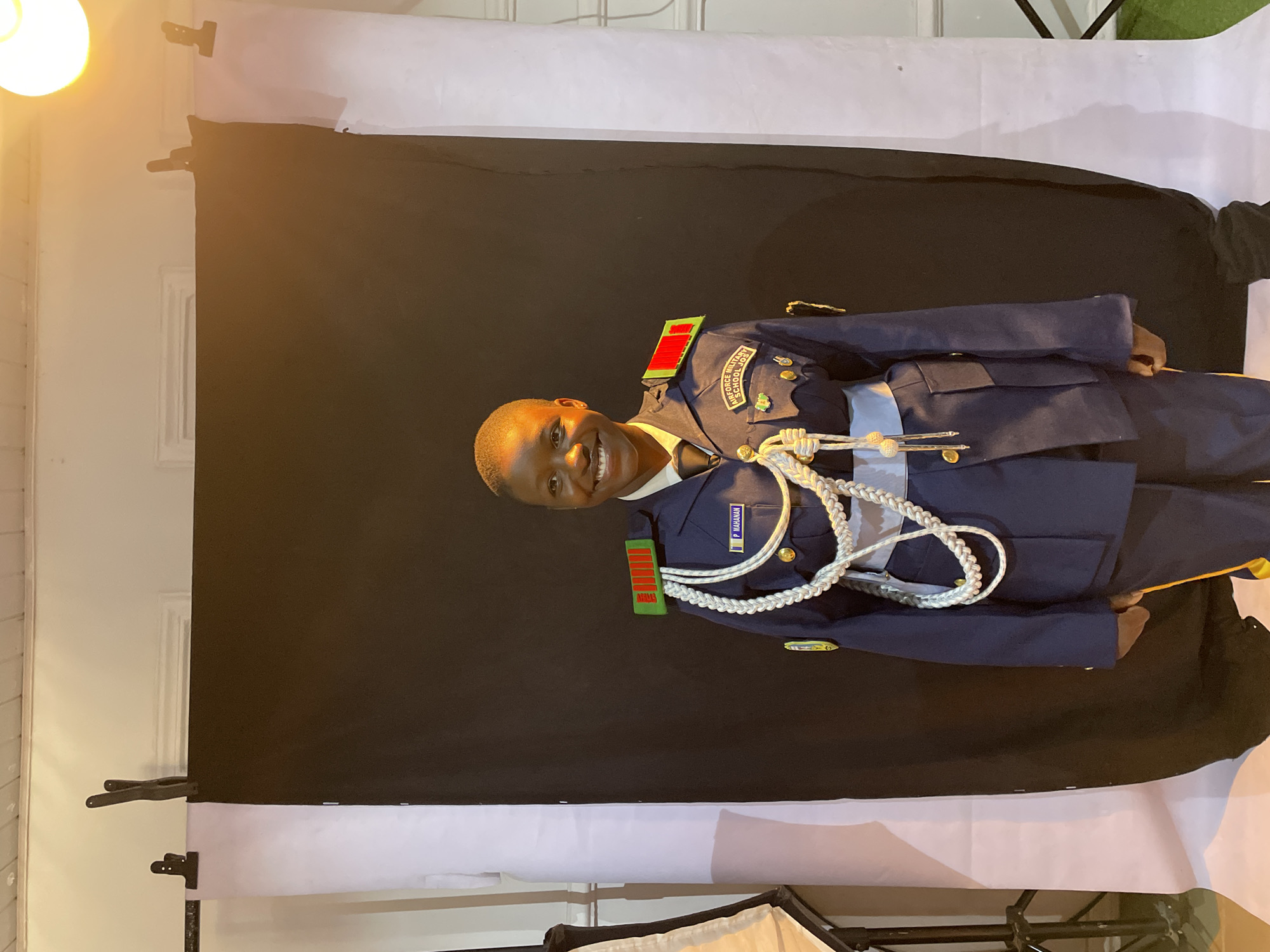The photograph depicts a young person in an Air Force Military School uniform standing in front of a backdrop consisting of black fabric over a white sheet. The uniform is blue with red and green epaulets, gold buttons, and a white rope cord across the chest. The name tag "P. MAHAMAN" is visible on the right chest. The background shows clamps holding the backdrop in place. The lighting is uneven, casting shadows on the backdrop. The location of Barakin Akawo, Nigeria is unknown and could not be confirmed.