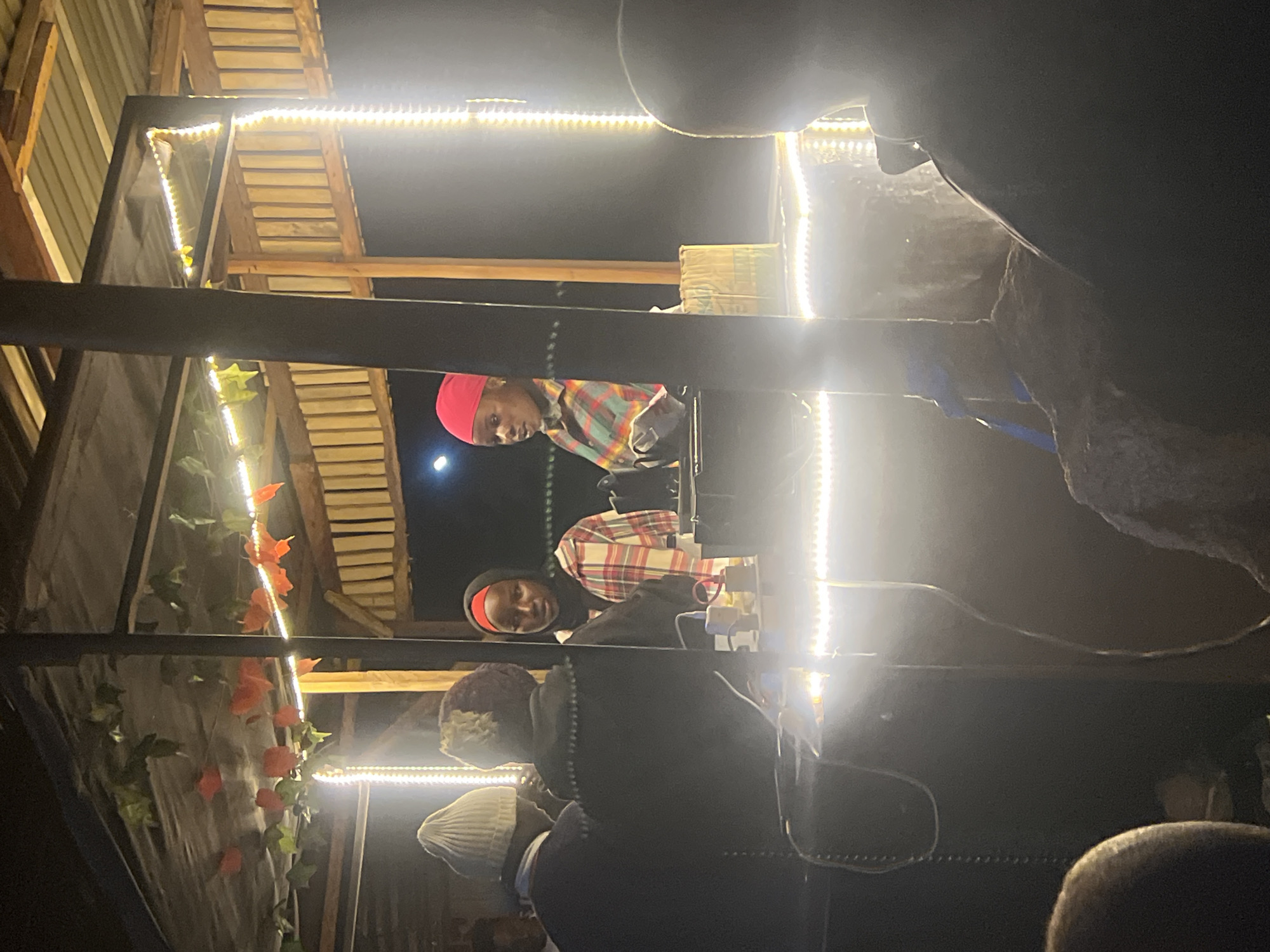 In Durumi, Nigeria, an outdoor food stall is illuminated by string lights. Two women, possibly vendors, are visible behind a counter. One woman wears a red headscarf and a plaid shirt, while the other has a black hijab. Several people, facing away from the camera, are seated or standing in the foreground, appearing to be customers. The stall has a wooden structure with a corrugated metal roof, decorated with artificial foliage and orange flower-like ornaments. Electrical wires and lights are prominent throughout the scene. The overall ambiance is that of a casual nighttime gathering at a local eatery.