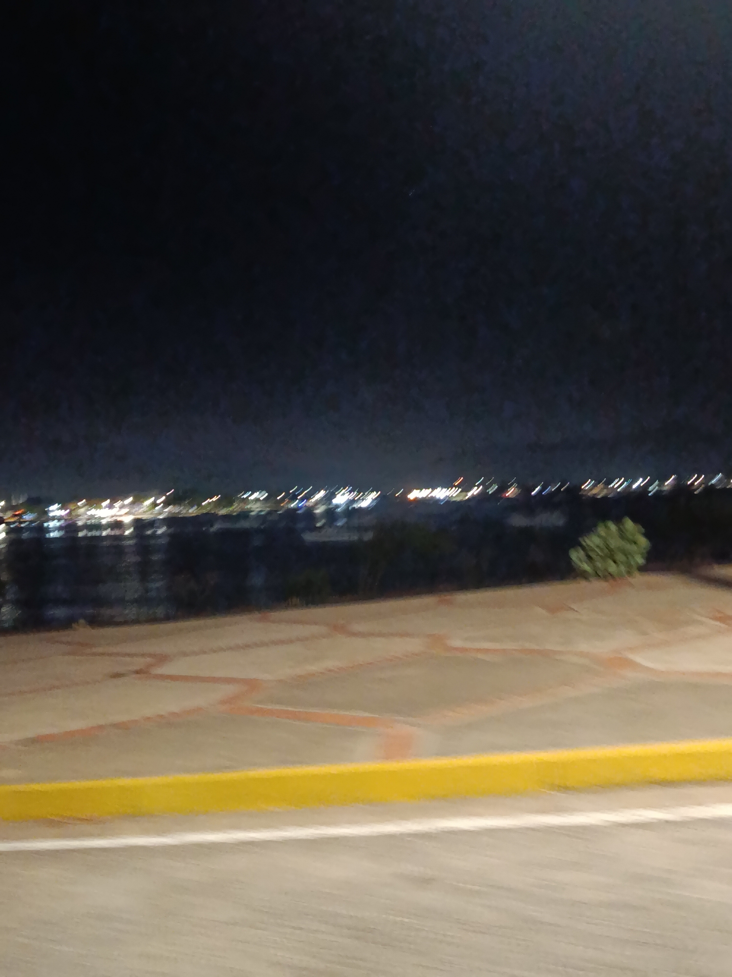 This image, likely taken at night in or near Juan Griego, Venezuela, depicts a scene from an elevated viewpoint. A dark sky covers the upper portion of the image. At the horizon line, there is a blurred, distant view of city lights reflecting on water. The middle ground features a paved area patterned with red/orange lines. A yellow curb separates this patterned area from a smoother gray paved surface in the foreground. No people are visible.