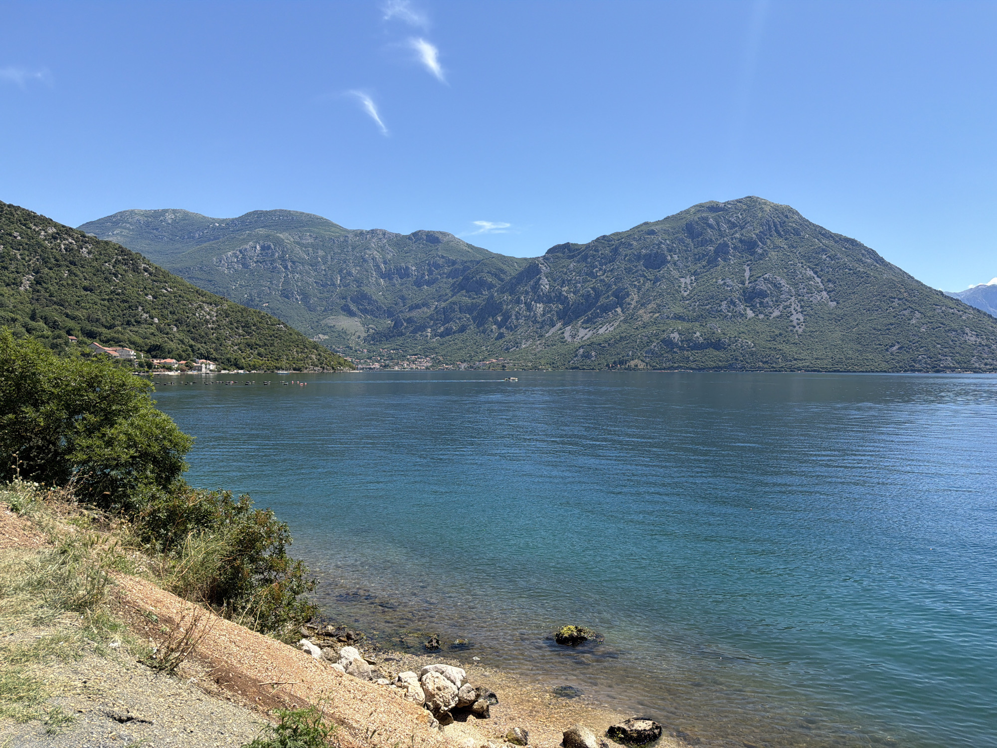 A bright, clear day is depicted in Lipci, Montenegro, with a vast expanse of blue water dominating the foreground and midground. The water is calm, with a turquoise hue near the shore, transitioning to a deeper blue further out. The shoreline is rocky and sandy, with some sparse vegetation and small bushes growing along the edge.
In the distance, a series of majestic, green-covered mountains rise steeply from the water's edge. The mountains are lush with vegetation, and their slopes are rugged and textured. Along the base of the mountains, a small, densely packed town or village can be seen, with buildings clustered together. Several small boats are visible on the water, some appearing to be moving.
The sky above is a brilliant, cloudless blue, with only a few wisps of white clouds scattered across the upper portion. The overall scene is one of natural beauty and tranquility, showcasing the striking landscape of the Montenegrin coast.