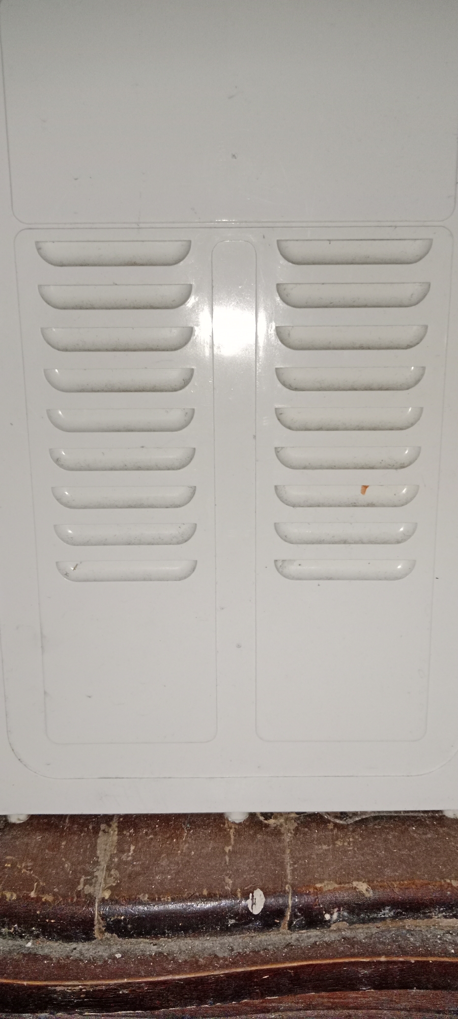 The image shows a close-up of a white plastic ventilation grill, which appears to be on an appliance or part of a building structure. The grill has two vertical sections, each containing seven horizontal, curved slats. There is some dust accumulated on the slats. Below the white grill is a dark, textured surface that resembles aged wood with visible grain and some wear. There are no people or specific landmarks visible that would allow for a precise location within Subang, Indonesia. The scene is static and appears to be an indoor or sheltered outdoor setting based on the materials.