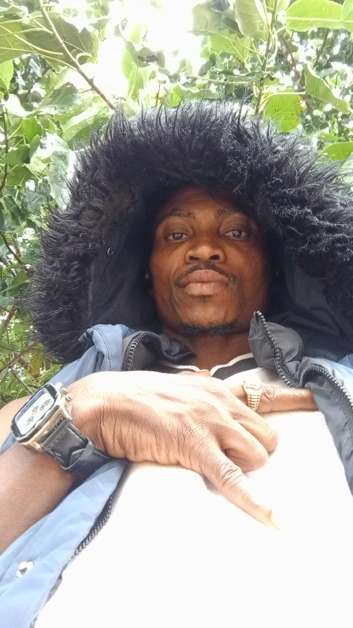 A close-up, over-the-shoulder selfie shows a Black male with dark skin and a mustache and goatee, wearing a blue jacket with a black fur-lined hood and a white shirt with black stripes. He is outdoors, with green leaves and branches of trees visible in the background, suggesting a natural or park setting. The man is looking directly at the camera with his mouth slightly open, and his right hand is resting on his chest, displaying a gold ring on his ring finger and a watch on his left wrist. The lighting appears to be natural sunlight, with some overexposure in the upper part of the image. The overall scene is casual and personal. No specific identifiable landmarks or details of Zango-ndaji, Nigeria are discernible from the image itself.
