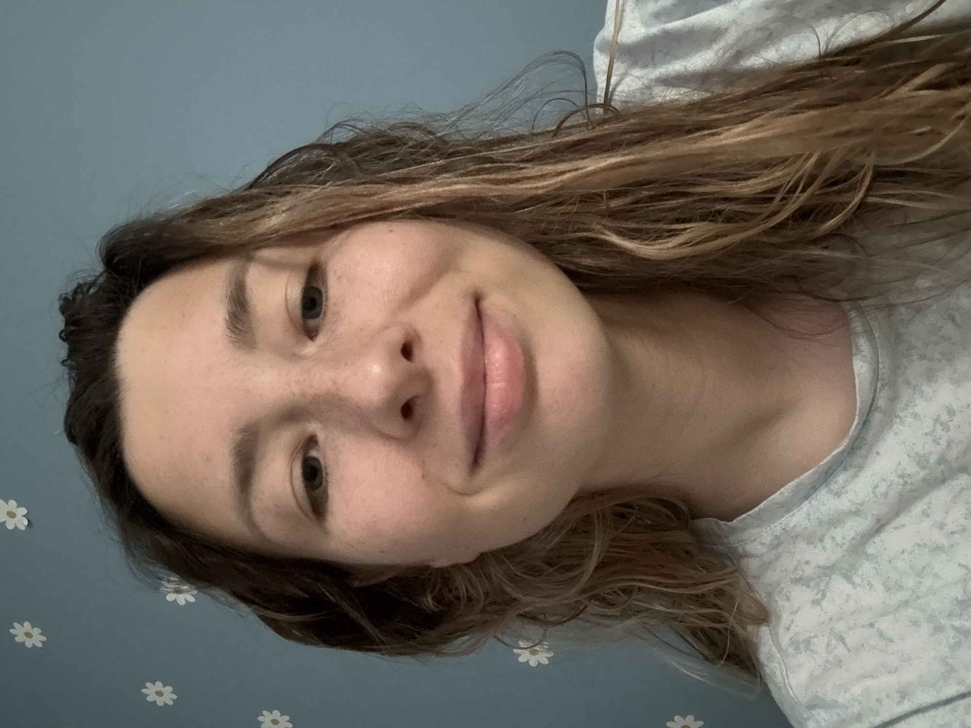 This is a close-up, eye-level, front-facing selfie of a young Caucasian woman with brown hair and light skin with freckles. She is wearing a light blue, patterned t-shirt. Her hair is damp and falls around her shoulders. The background is a light blue wall with small white flower decals. The image does not provide any information or context that would allow for determining a specific location in Moscow, Russia.