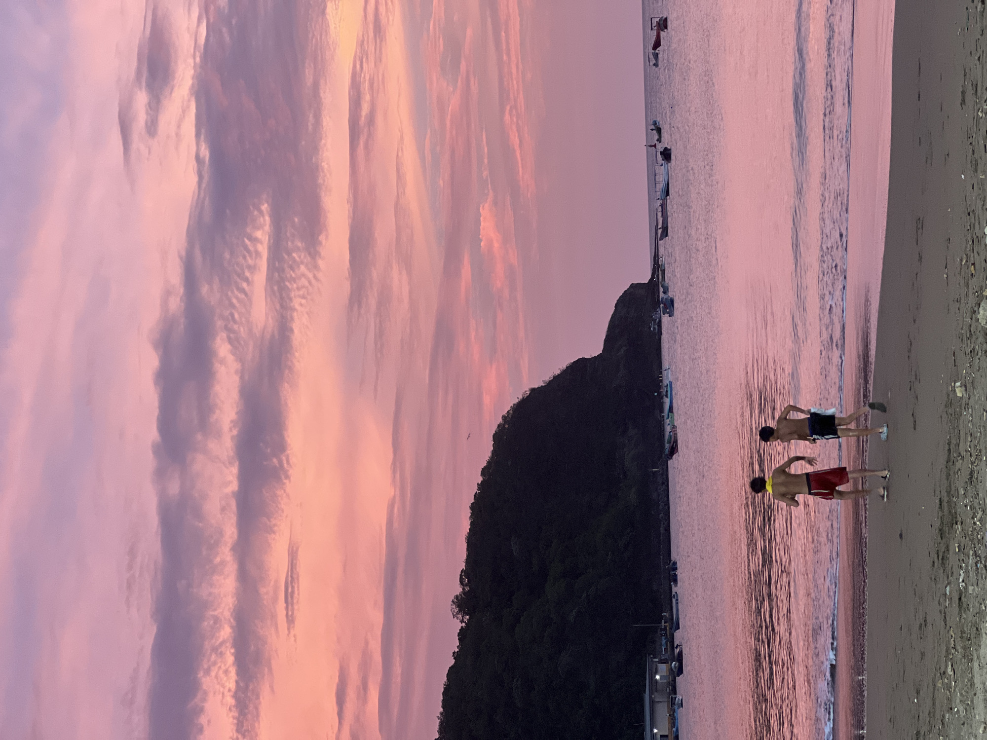 The image depicts a serene sunset scene at the beach in San Juan del Sur, Nicaragua. Two young males are walking along the sandy shore, facing away from the camera. They are wearing swim trunks, and one has a yellow band around his neck. The ocean is calm with gentle waves lapping at the shore, reflecting the vibrant pink and purple hues of the sky. In the distance, a tree-covered hill juts out into the water, casting a dark silhouette against the colorful sky. Several small boats are anchored in the bay, adding to the tranquil atmosphere. The sky is filled with soft, wispy clouds painted in shades of pink, lavender, and orange, characteristic of a tropical sunset. The overall impression is one of peacefulness and natural beauty.