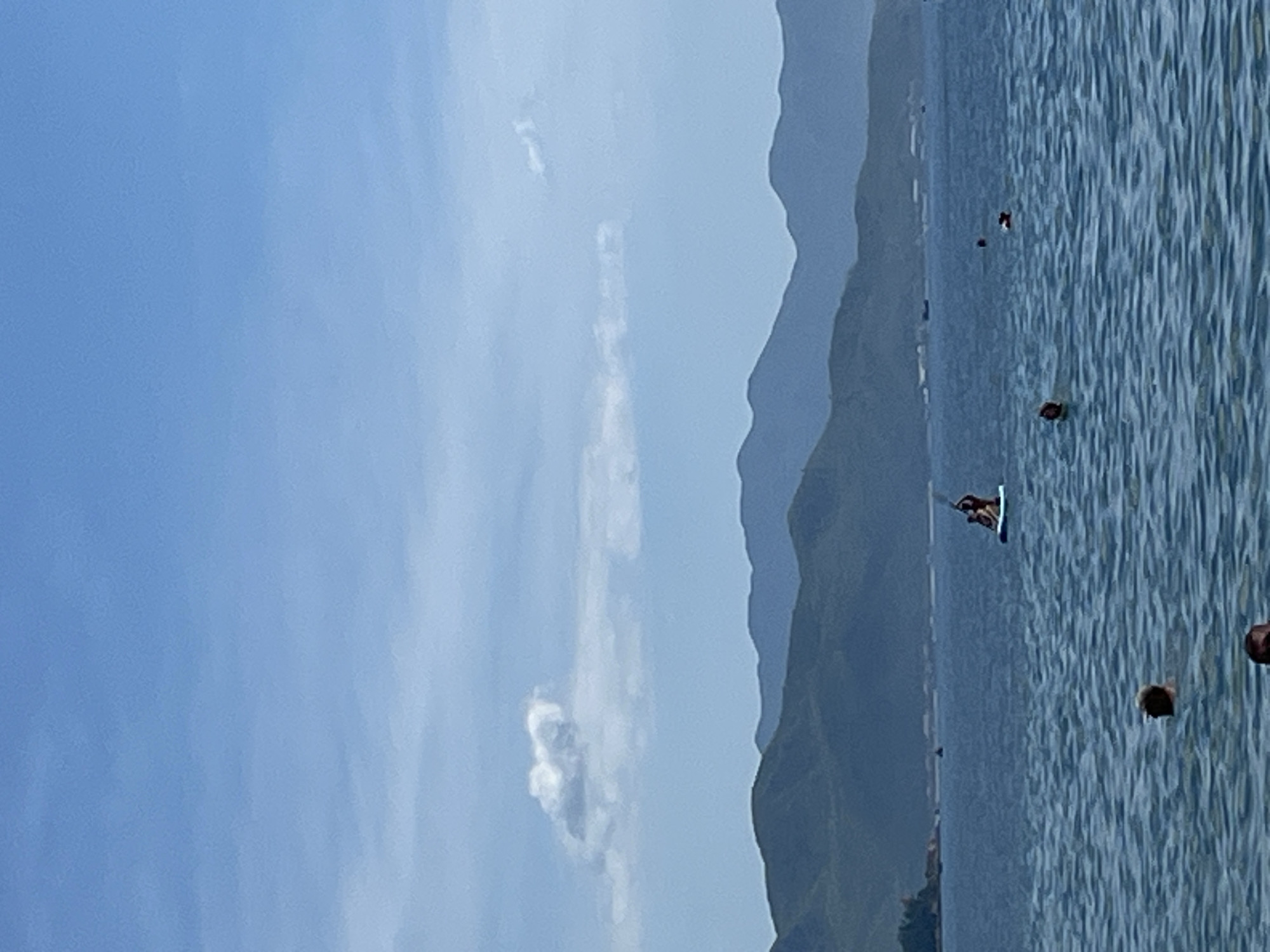 This is a vertical, outdoor, medium-shot photograph taken on a sunny day in Nha Trang, Vietnam. The image captures a serene seascape with layered, hazy green mountains in the background, meeting a clear blue sky dotted with wispy white clouds. The foreground is dominated by a calm, blue ocean with gentle ripples.
In the mid-ground of the water, two people are visible on a paddleboard. One person stands and appears to be paddling, while another person is seated behind them. Further out in the water, several other individuals are swimming, with only their heads visible. One person is in the foreground, also swimming.
The coastline is visible on the far left, featuring some vegetation and buildings, suggesting a populated area along the shore. A few small boats are also present in the distance. The overall scene conveys a tranquil atmosphere of a recreational day at the beach.